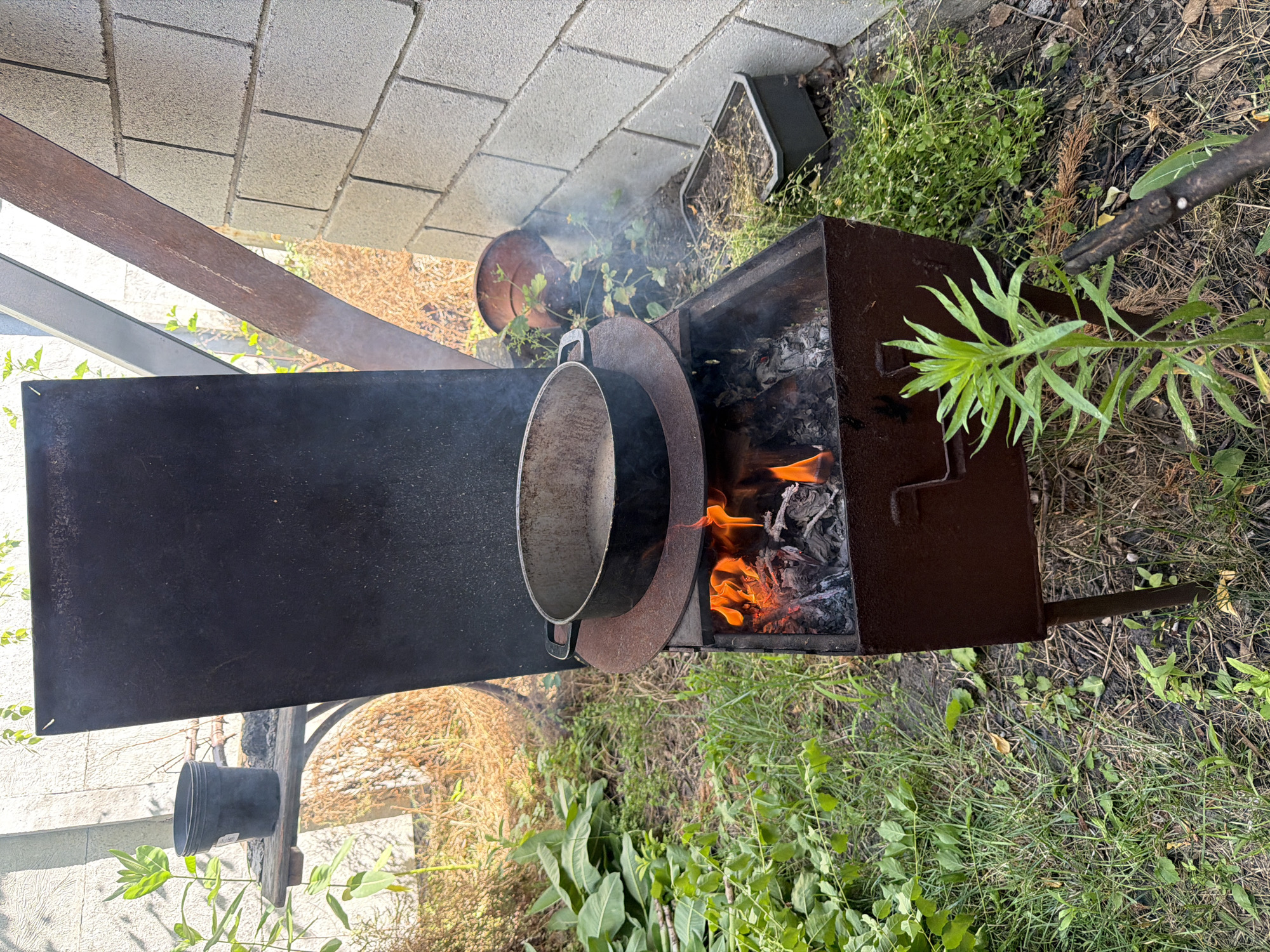 The image depicts an outdoor scene with a metal barbecue grill actively burning a fire. A large metal pot is placed on top of the grill, suggesting cooking is in progress. The grill itself is situated on the ground amidst lush green grass and various plants, with some dry leaves and debris scattered around. In the background, there's a wall constructed from grey concrete blocks. To the left of the grill, a long, narrow piece of rusted metal is visible, possibly part of a structure or support. A black plastic pot is hanging from a wooden pole. The overall setting appears to be a backyard or garden area. The lighting suggests it is daytime, and the weather seems to be fair, with no indication of rain. There is no visible text in the image.