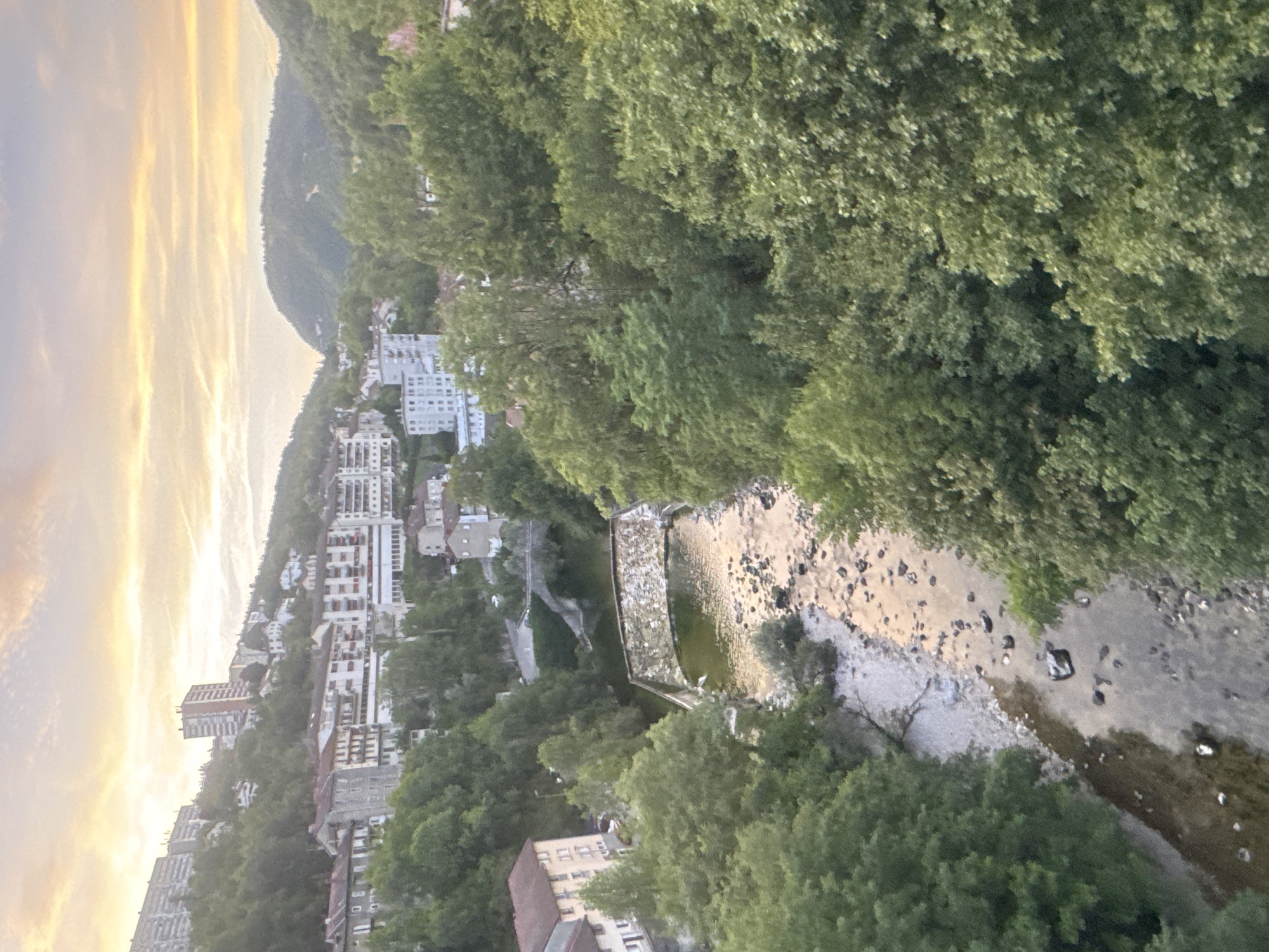 This image captures a scenic view of the city of Saint-Claude, France, at dusk. The foreground is dominated by a winding river, its surface reflecting the warm, golden hues of the setting sun. The riverbed is visible in places, strewn with pebbles and rocks. Lush green trees line the banks, their dense foliage creating a natural frame for the scene.
In the middle ground, a stone bridge arches over the river, connecting two sides of the town. Beyond the bridge, more trees and vegetation give way to a hillside dotted with buildings. These structures, varying in size and architectural style, are characteristic of a European town nestled in a valley. Some appear to be apartment complexes with multiple stories, while others are smaller, more traditional houses.
The background reveals a dramatic landscape of rolling hills and mountains, softened by the twilight glow. The sky above is a canvas of soft, pastel colors, with streaks of orange, yellow, and pink clouds, indicating the transition from day to night. The overall impression is one of tranquility and natural beauty, with human habitation integrated harmoniously into the environment. The scene is devoid of any visible people or specific actions, presenting a static yet evocative snapshot of Saint-Claude.