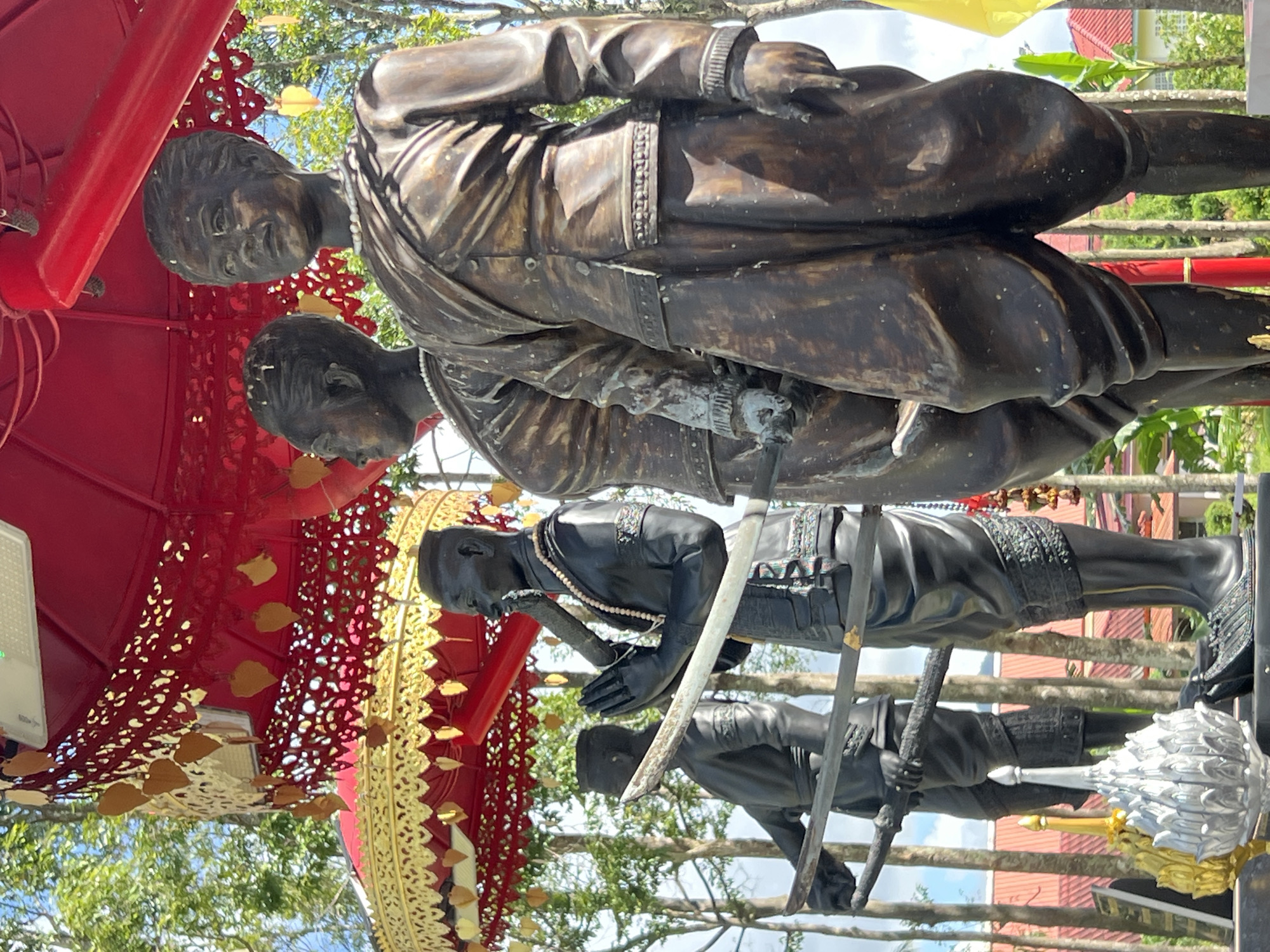 In Thep Krasatti, Thailand, a statue of four figures stands under a red and gold umbrella. The two figures in the foreground are made of bronze and wear traditional clothing. The figure on the right has a pearl necklace and wears a uniform with a sash. The figure to its left also wears a uniform and a pearl necklace. Behind these two figures, two more statues are visible. The statue on the left is black and depicts a figure with praying hands and a pipe-like object in its mouth, also wearing a pearl necklace. The statue furthest to the left is also black and holds a sword. The statues are positioned in front of trees and buildings with red roofs.