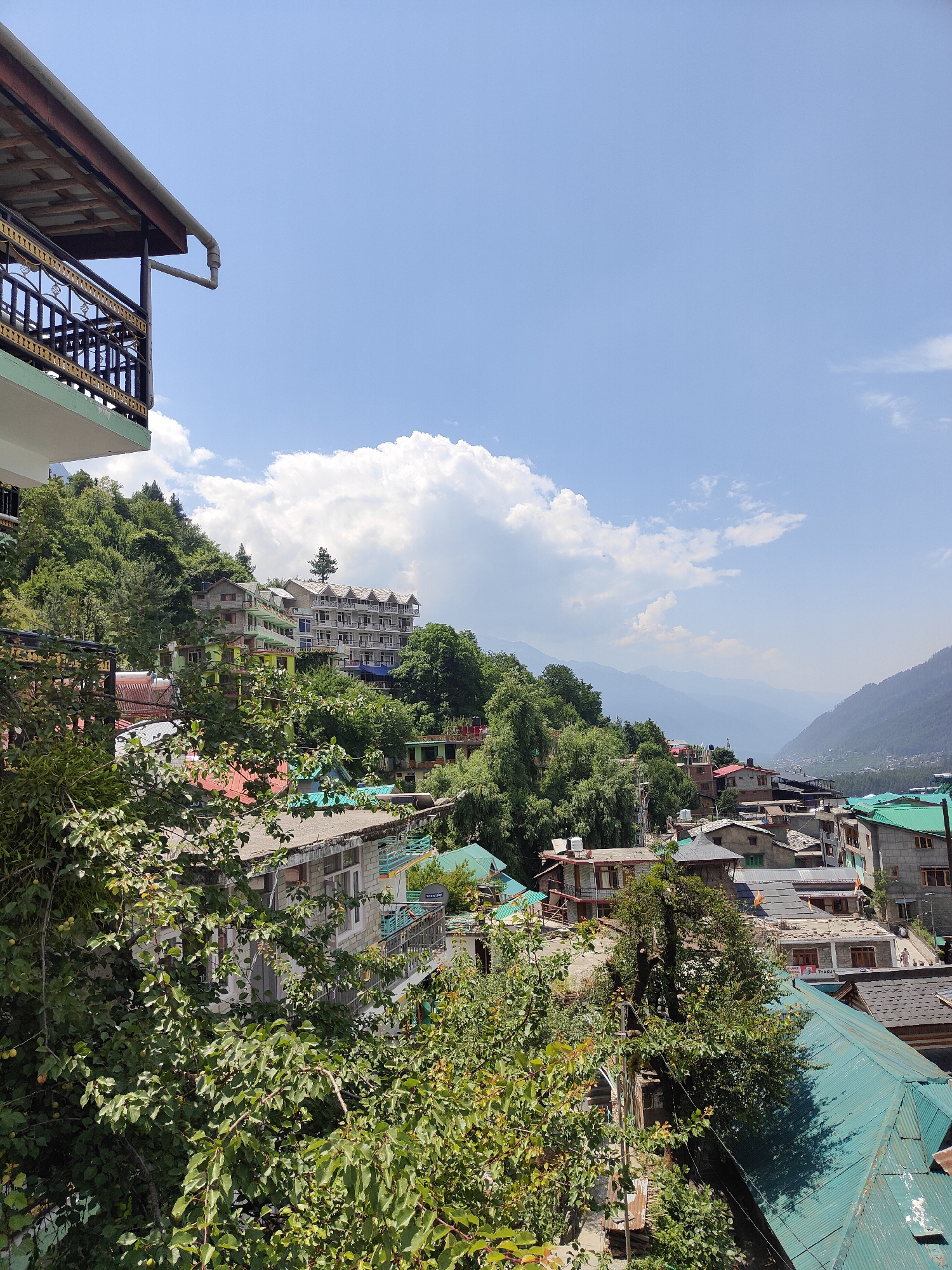 This image captures a scenic view of a hillside village in Vashisht, India. The foreground is dominated by lush green foliage, with branches and leaves partially obscuring the view of buildings below. Several houses and buildings, constructed with a mix of concrete and stone, are visible on the terraced slopes. These structures feature a variety of roof types, including corrugated metal in shades of green and grey, and traditional tile roofs.
A prominent multi-story building with a white facade and green balconies stands out on the hillside, surrounded by trees. Further in the background, a sprawling hotel-like structure with multiple floors and a distinctive patterned roof is nestled against the lush, tree-covered mountain. The buildings cascade down the slope, creating a dense village environment.
In the distance, layers of mountains recede into a hazy blue, suggesting a vast valley. The sky is a vibrant blue, dotted with fluffy white clouds, indicating a clear and sunny day. The lighting suggests it is daytime, likely late morning or afternoon, with bright sunlight casting shadows. There are no visible people in the image, and the scene conveys a sense of tranquility and the natural beauty of the mountainous region. A sign with the text "THAKUR" is visible on one of the buildings in the middle ground.