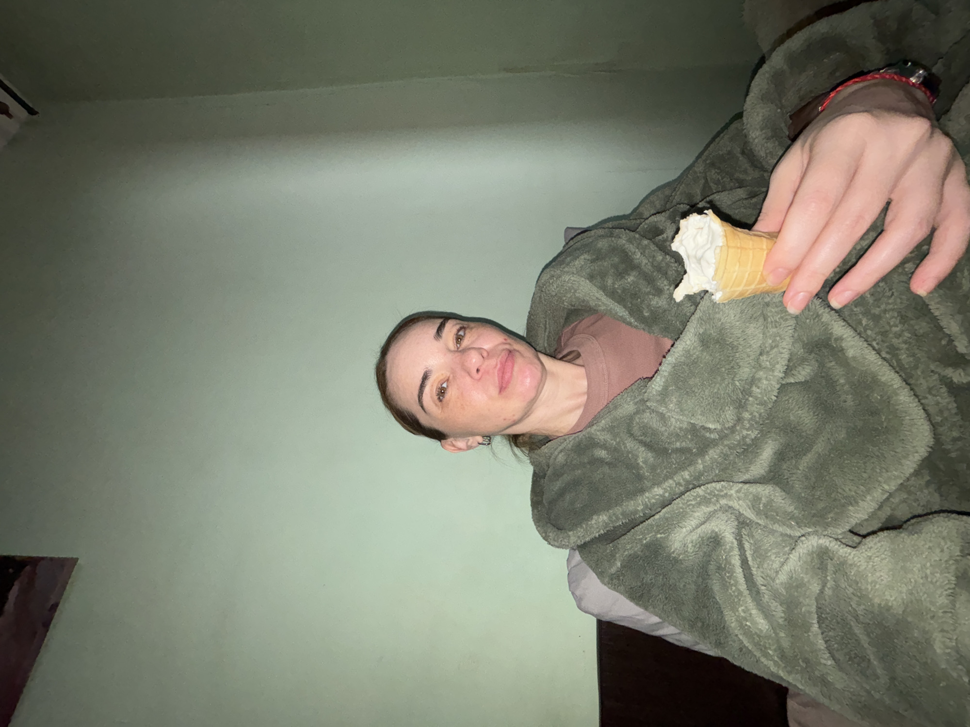 A woman is lying in bed in a room with light green walls, covered in a fuzzy green blanket. She is holding a partially eaten ice cream cone in her right hand. She wears a brown top. The location is Unknown, Unknown.