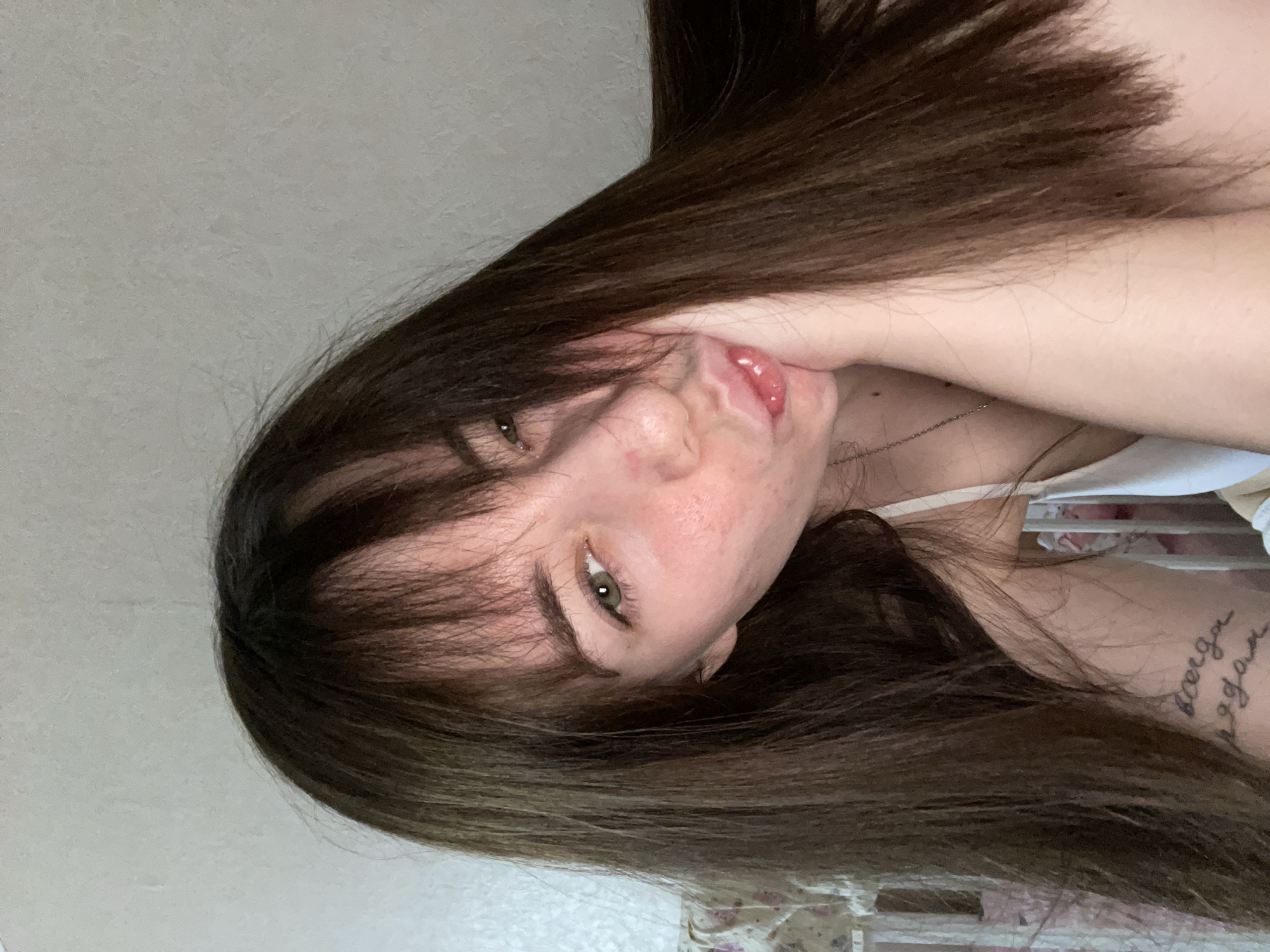 A selfie of a woman with brown hair is oriented at an approximate 90-degree angle. She is positioned indoors, facing the camera with her head tilted. Her right hand is raised, partially obscuring her neck and jawline. She has a neutral to slightly pouting facial expression. A tattoo is visible on her upper left arm. The background includes a plain wall. The exact location within the city of Chaikovsky, Russia cannot be determined from the image.