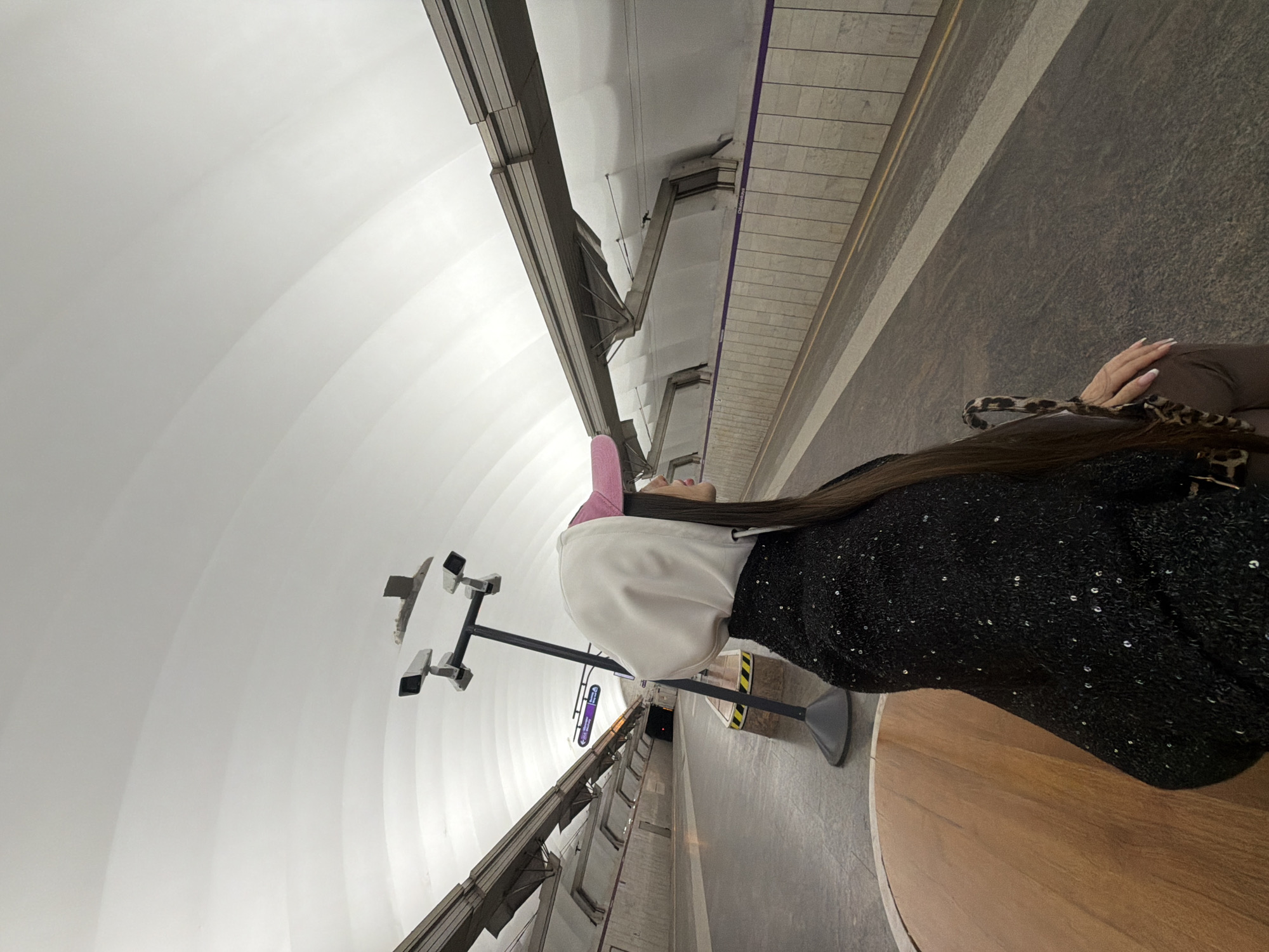 A long shot shows a woman sitting on a circular wooden bench in what appears to be a metro station, likely in Shushary, Russia, based on the station design. The station features a white, arched ceiling. The woman is wearing a white hooded jacket, a pink hat, and a sparkly black dress. She has long, dark hair and is holding a brown bag on her lap. The metro station has a modern design, with security cameras mounted on poles and tracks along the sides of the platform. The platform is grey.