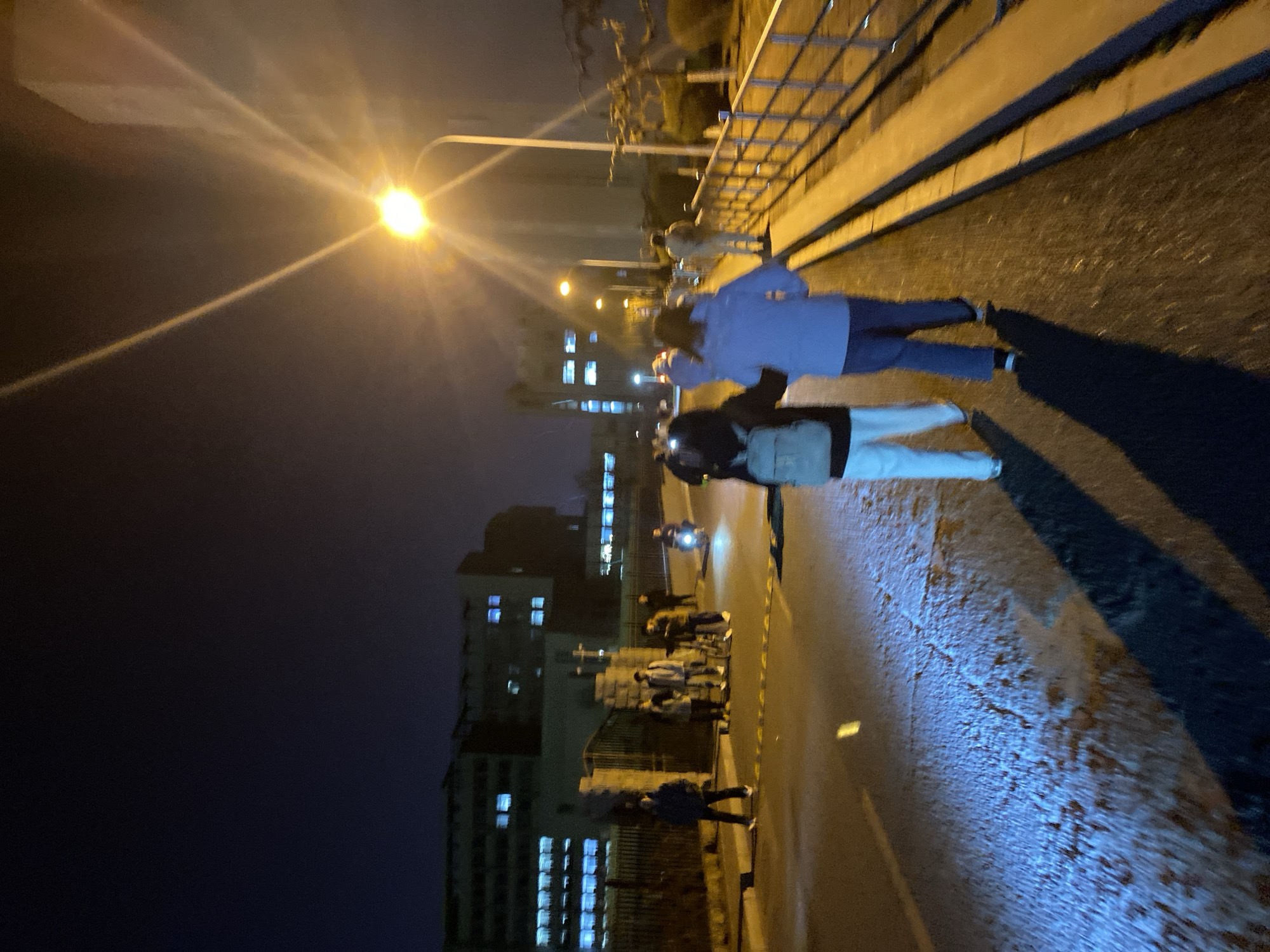 Night scene along a street in Zhongtun, China. A bright street lamp illuminates the right side of the image, casting long shadows. Two people walk on the sidewalk, one wearing a light blue shirt and the other a dark jacket with light-colored pants. Further down the street, a group of people stand near a barrier. A motorcyclist is visible. Buildings with lit windows line the left side of the street, contrasting with the dark sky.