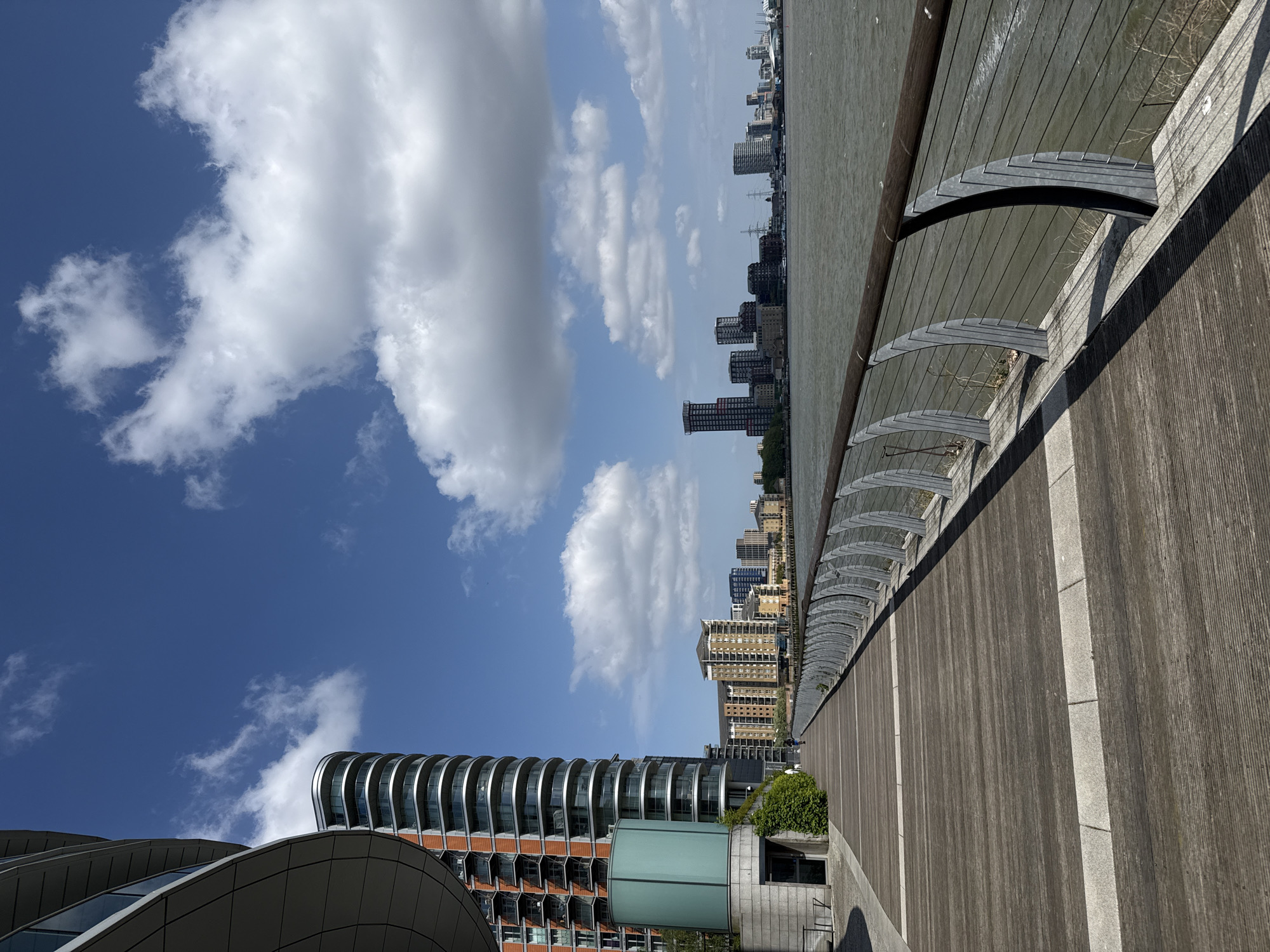 This image depicts a modern urban waterfront scene on a bright, sunny day, under a clear blue sky with scattered white cumulus clouds. The photograph is oriented vertically, showing the horizon diagonally.
In the immediate foreground and extending to the right, a wide wooden promenade or boardwalk is visible, bordered by a textured concrete wall and a contemporary railing system. This railing features multiple arched metal supports with taut vertical wires or cables, creating an architectural detail along the water's edge.
To the left, a distinctive, multi-story building with modern architecture stands prominently. It showcases a curvilinear design with tiered balconies or facade elements that create a ribbed, wave-like appearance. The building is constructed with reflective glass and reddish-brown cladding. At its base, a light-green, cylindrical structure is visible, surrounded by some vibrant green foliage.
Across the expanse of water, which appears calm and greyish-green, a densely packed cityscape stretches into the distance. Numerous high-rise buildings of varying heights and contemporary designs form the skyline on the opposite bank. Small details, possibly cranes or masts from boats, can be faintly discerned among the distant structures. A single, small, and somewhat blurry person is visible walking on the wooden promenade towards the middle of the frame.
The scene conveys a sense of peaceful urban development along a major waterway, likely during the daytime given the bright illumination and clear shadows. The architectural style of the foreground building and the overall setting strongly suggest a location within London, United Kingdom, specifically along the River Thames or a similar dockland area. There is no visible text in the image.