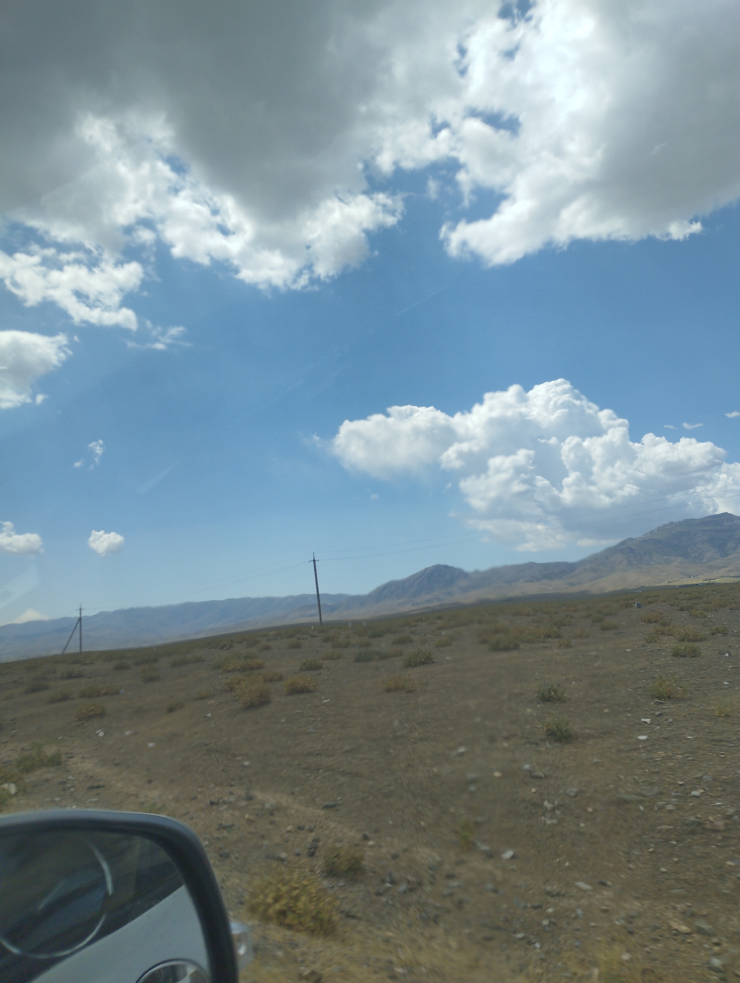 The image captures a vast, open landscape under a partly cloudy sky. The terrain is dry and covered with sparse vegetation, appearing as rolling hills and plains leading to distant mountains. Two utility poles with wires are visible, indicating some infrastructure in the area. In the foreground, a portion of a car's side mirror is visible, reflecting the sky and the interior of the vehicle. The overall scene suggests a remote or rural location, likely within the city of Forish QFY, Uzbekistan, characterized by its natural, undeveloped environment.