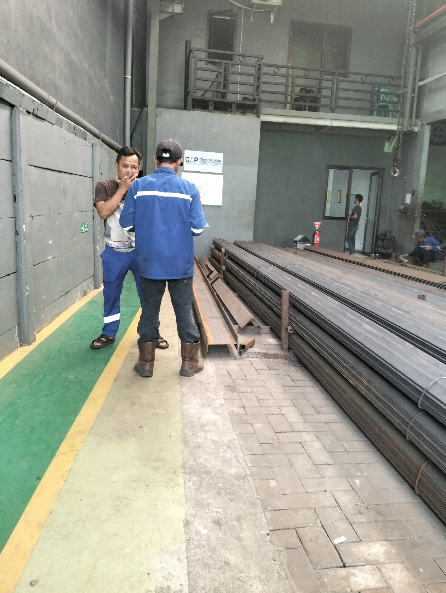 Two men are standing in what appears to be an industrial setting in Gunung Sindur, Indonesia. The man on the left, wearing a brown t-shirt and blue pants with white stripes, is speaking to the man on the right. The man on the right is wearing a blue jacket with white stripes and dark pants, and a black baseball cap. He is facing away from the camera, with his back to the viewer. In the background, stacks of metal are visible. A green floor with a yellow line runs along the left side of the image, and a brick walkway is on the right. A red fire extinguisher is mounted on the wall behind the men.