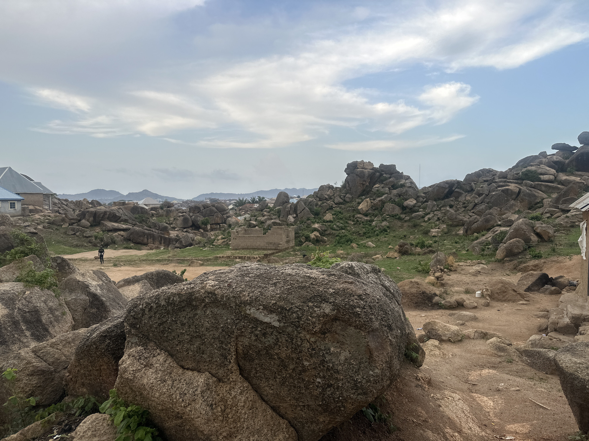 The image depicts a rocky landscape in Jos, Nigeria. Large granite boulders dominate the foreground and midground, with scattered vegetation amongst the rocks. In the distance, residential buildings are visible. A single person is walking in the midground. A partially constructed brick structure can be seen in the middle distance. The sky is overcast with scattered clouds.