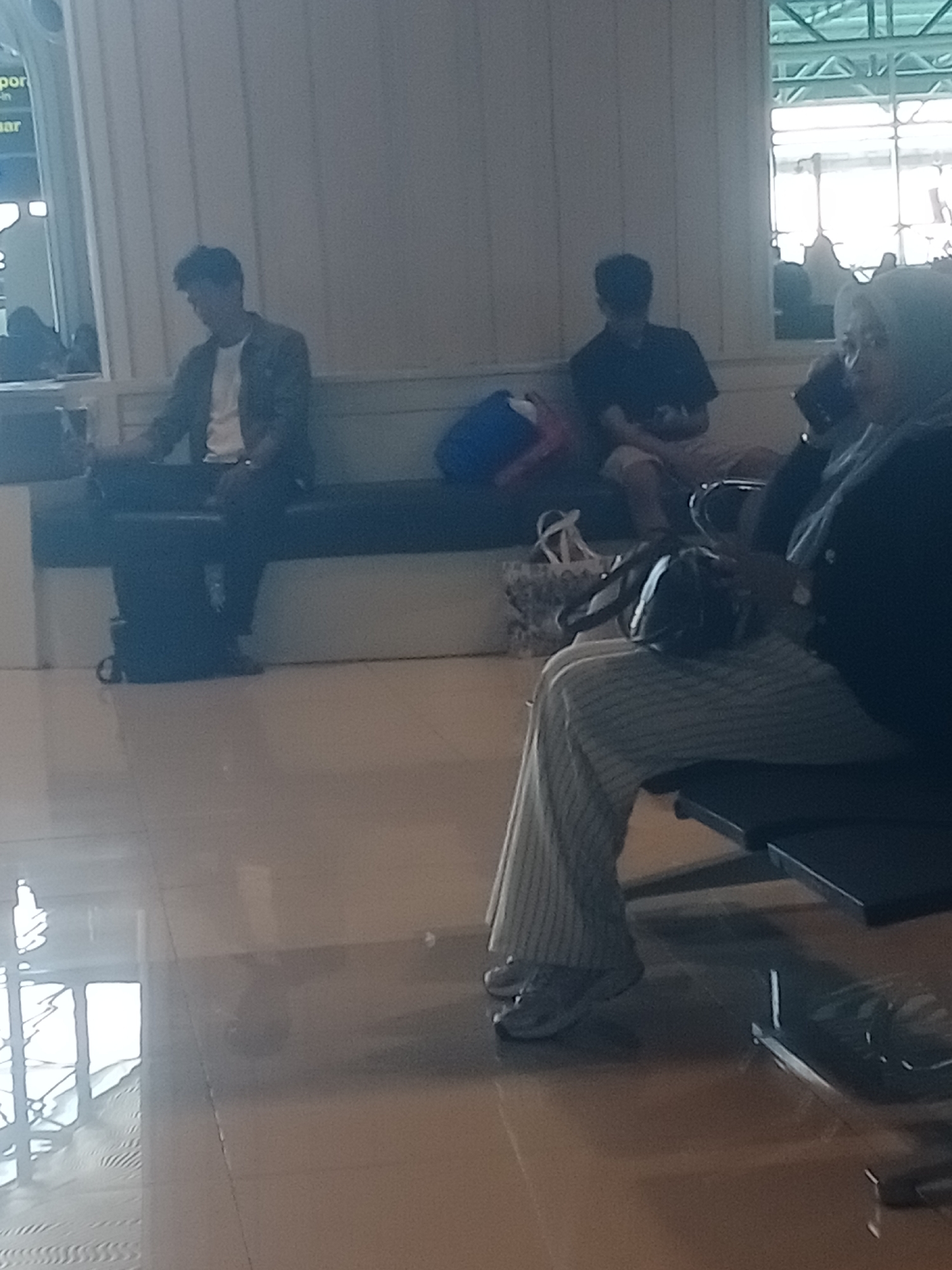 The image captures a scene inside what appears to be a waiting area, possibly a ferry terminal, in Baji Mangngai, Indonesia. In the foreground, a person wearing a hijab and a dark jacket with striped pants is seated, holding a dark object, likely a phone, to their ear. To their left, another person in a dark shirt and light-colored shorts is seated, looking down at their hands. Further to the left, a third person, a male wearing a light-colored t-shirt under a dark jacket and dark pants, is seated, looking downwards. Adjacent to him, a dark backpack rests on the floor. The environment features a long, dark bench where the individuals are seated. The background shows a wall and a partially visible window structure with a green tinted roof. The floor is a polished, light-colored surface reflecting the ambient light.
