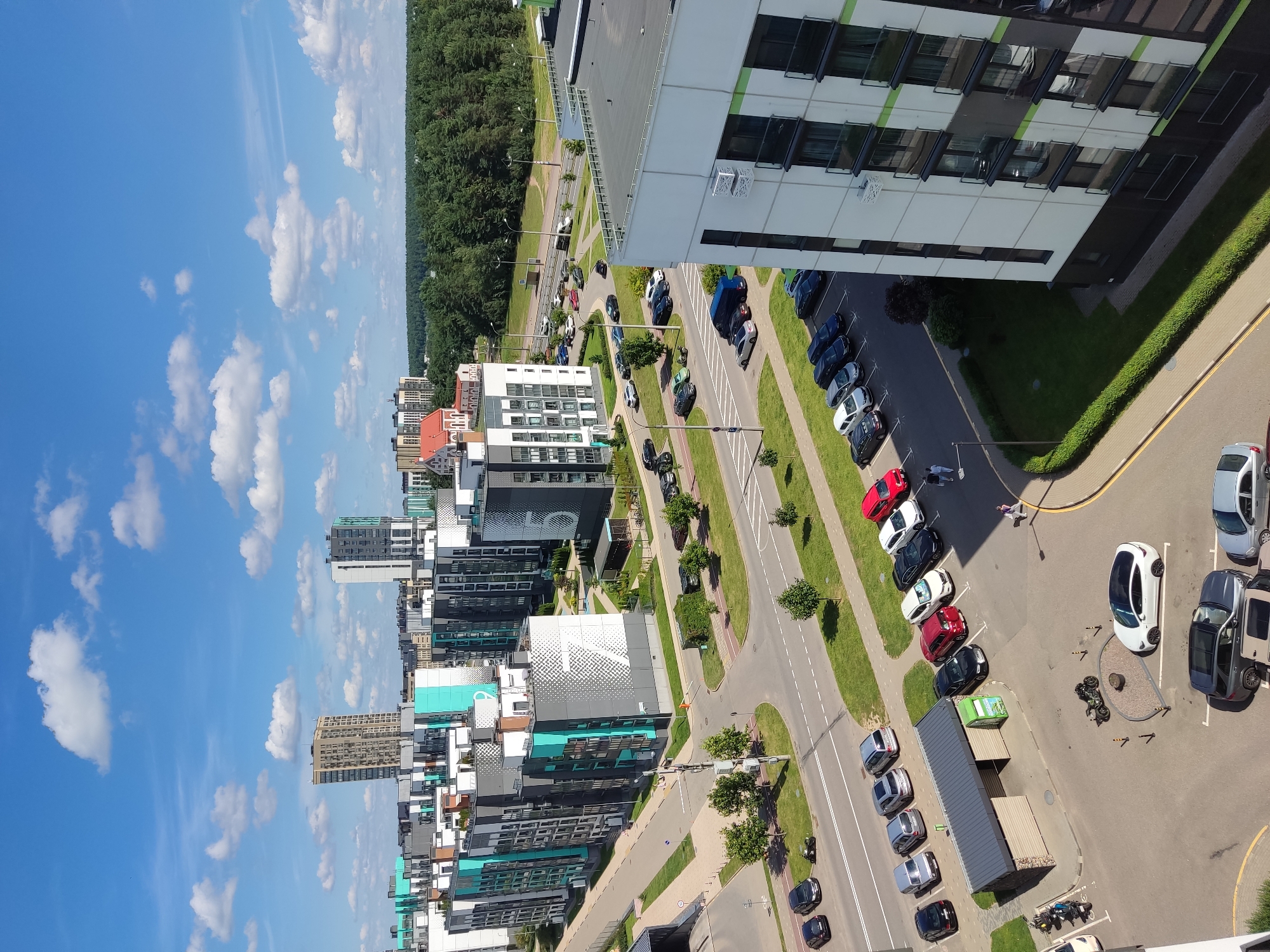 The aerial photograph, captured on a bright, sunny day with scattered white clouds against a blue sky, depicts a modern residential area in Бараўлянскі сельскі Савет, Belarus. The scene is dominated by multi-story apartment buildings with a contemporary design, featuring a mix of grey, white, and turquoise facades. Several of these buildings are marked with large white numbers, including a '7' and a '5' on prominent structures.
In the foreground, a white Tesla Model S is parked in a paved lot. Adjacent to it is a dark grey SUV and a silver sedan. Further into the scene, a row of parked cars lines a street next to a covered bus stop. These vehicles include a black SUV, a red sedan, a white sedan, a black sedan, and a white hatchback. There are also two motorcycles parked near a small, covered structure.
The apartment complexes are interspersed with well-maintained green lawns and small trees. Several streets and pathways are visible, with cars navigating the area. In the background, a dense forest stretches across the horizon, with taller, older buildings visible further in the distance. The overall impression is one of a well-developed urban or suburban landscape, likely a new housing development.