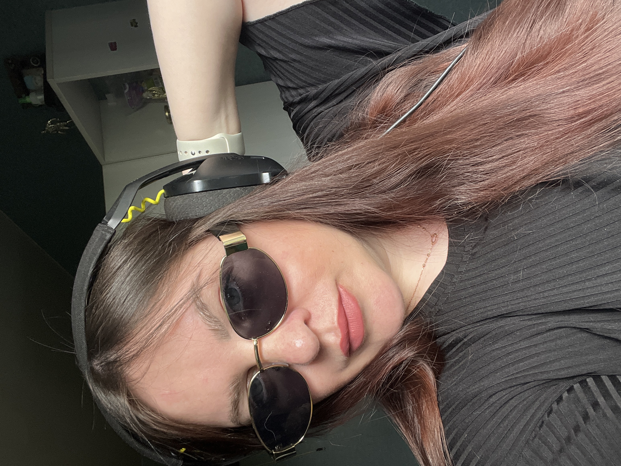 A medium shot depicts a young woman with brown hair wearing black-rimmed sunglasses and a black ribbed top. She is wearing headphones and an Apple Watch on her left wrist. The background includes a white cabinet. The city and location of Noviy, Russia is not discernible from the information provided.