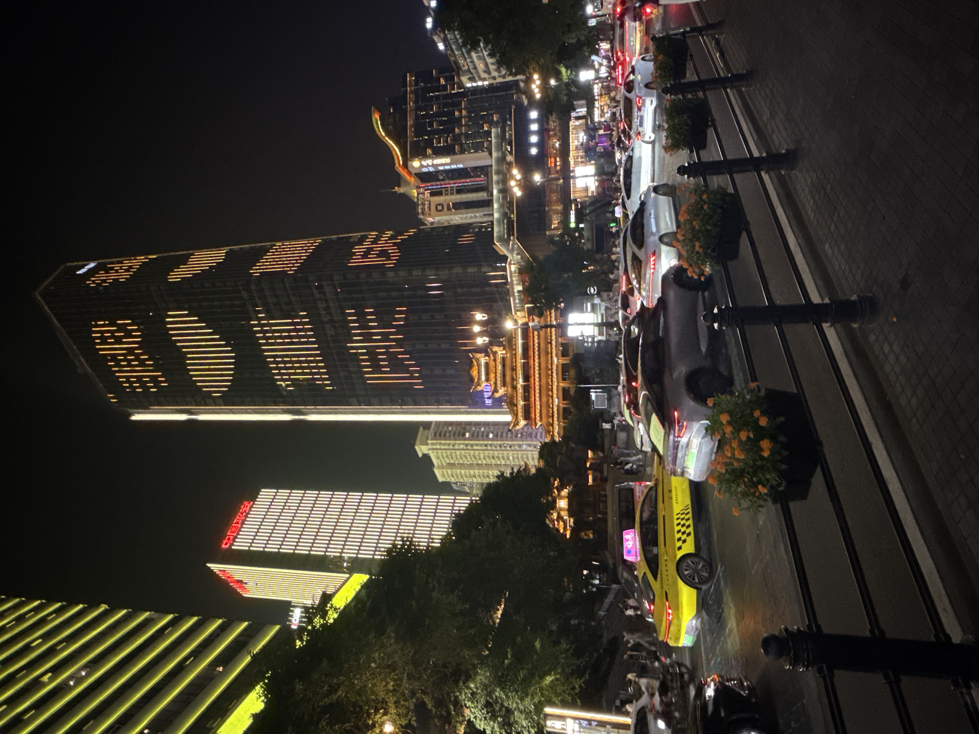 This image depicts a nighttime street scene in the Yuzhong District of Chongqing, China, characterized by illuminated skyscrapers, traffic, and pedestrians. Dominating the background is a very tall, dark skyscraper with a distinctive pattern of vertical orange lights, possibly forming letters or symbols. To its left, another skyscraper is adorned with vertical yellow lights. In the middle ground, a traditional Chinese-style building with a curved roof, also lit up, sits below other modern buildings. The street in the foreground is filled with vehicles, including a bright yellow taxi with black checkered patterns on its side and a pink advertisement on its roof. Several other cars, including white sedans, are visible in traffic. A few pedestrians are walking on the sidewalks and crossing the street. The foreground features a black metal railing with planters containing orange flowers. The overall impression is a busy urban environment at night, with a blend of modern architecture and traditional elements.