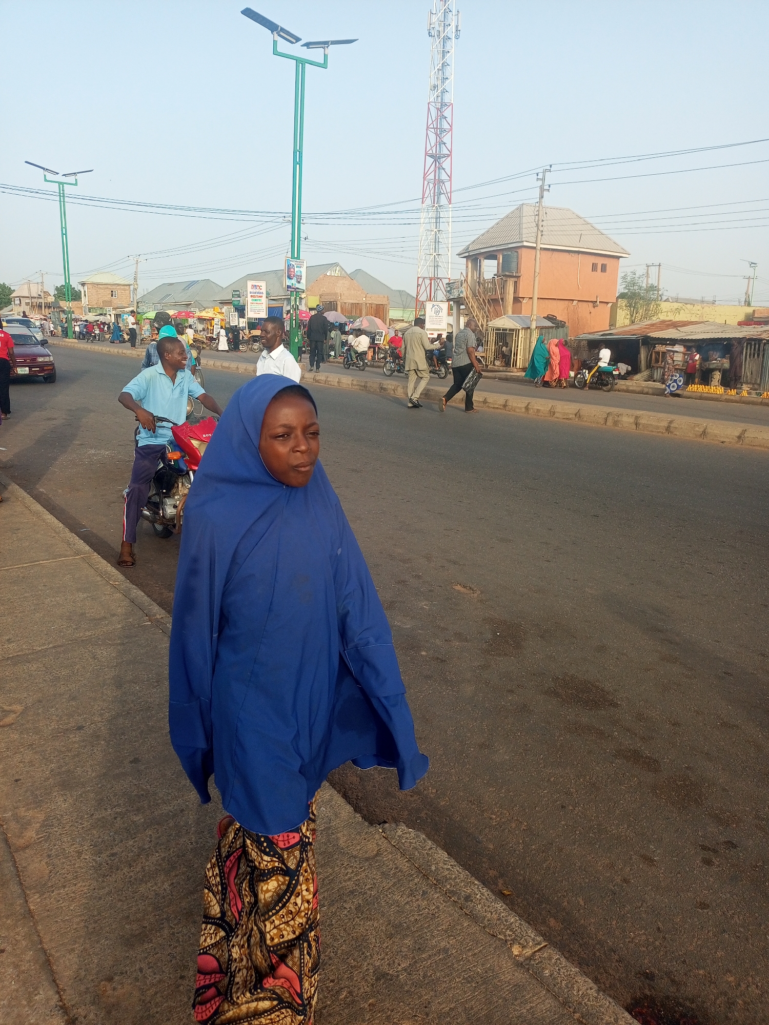 A woman wearing a blue head covering and a patterned skirt walks along the edge of a road in Tempure, Nigeria. Behind her, a man sits on a motorcycle. Other people are visible on the sidewalks and in the background, along with buildings, streetlights, and a telecommunications tower. Shops and market stalls line the sidewalks.