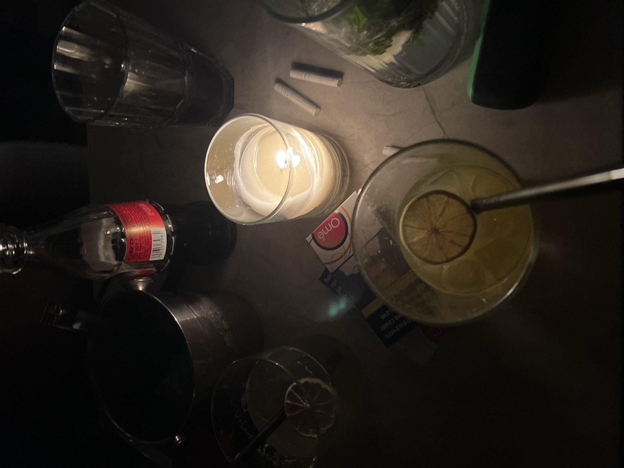 The scene appears to be a dimly lit tabletop, possibly in a bar or lounge in Plovdiv, Bulgaria, given the "One" brand cigarette pack present, a brand sold in Bulgaria. Several objects are scattered across the surface. There are two glasses containing liquid, possibly cocktails, one with a lime garnish and a straw. A clear glass candle holder contains a lit candle. There are two cigarettes next to it. A bottle of Coke is partially visible. A metallic cocktail shaker is present. A tall, empty glass is near the upper left. Overall, the scene evokes a relaxed and dimly lit ambiance.