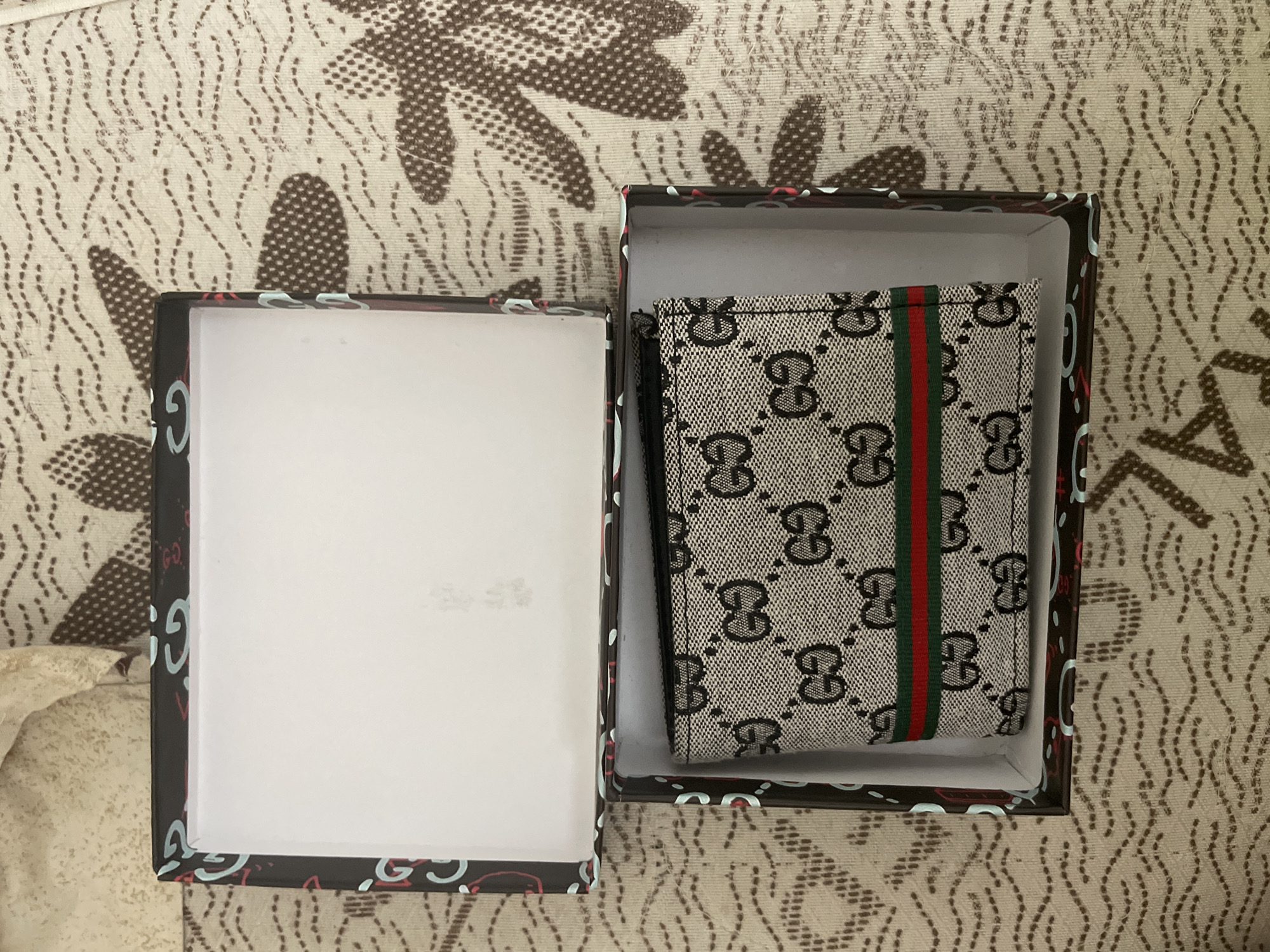 An overhead photograph captures a beige and black "GG" monogram patterned wallet, featuring a distinct red and green vertical stripe, resting inside an open box. The wallet is positioned on the right side of the image, contained within one half of the dark-colored box. The interior of the box is white, while its exterior edges display a repeating pattern of stylized light blue and red "Gucci" or "GG" logos. The other half of the box, visible on the left, is empty, also featuring a white interior and similarly patterned exterior edges. Both the wallet and the box are placed on a light-colored fabric surface adorned with an abstract brown pattern, possibly floral or leaf-like, interspersed with faint wavy dotted lines. The arrangement suggests the display or unboxing of a new item, mimicking luxury brand aesthetics.