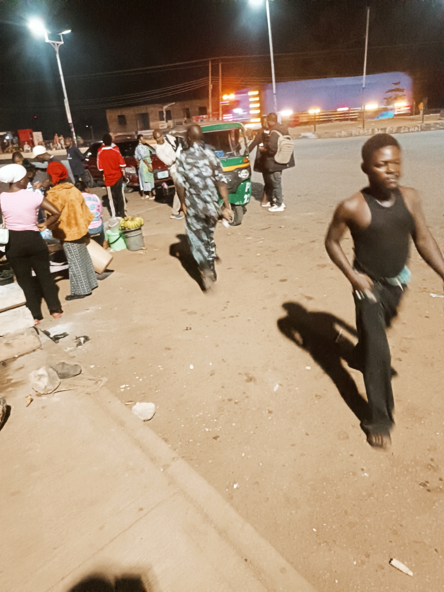 The image depicts a street scene in Bukuru, Nigeria, at night. Streetlights illuminate the scene. Several people are present, engaging in various activities. One man walks toward the viewer, wearing a black tank top and dark trousers, his feet bare. Other individuals are gathered near what appears to be a roadside vending setup, indicated by buckets and containers. A green and yellow auto rickshaw (tuk-tuk) is positioned nearby. In the background, a large truck with illuminated lights is visible on a road, indicating traffic. Buildings line the street in the distance. The ground is a mix of paved and unpaved surfaces. The overall atmosphere suggests a lively, informal commercial area at night.