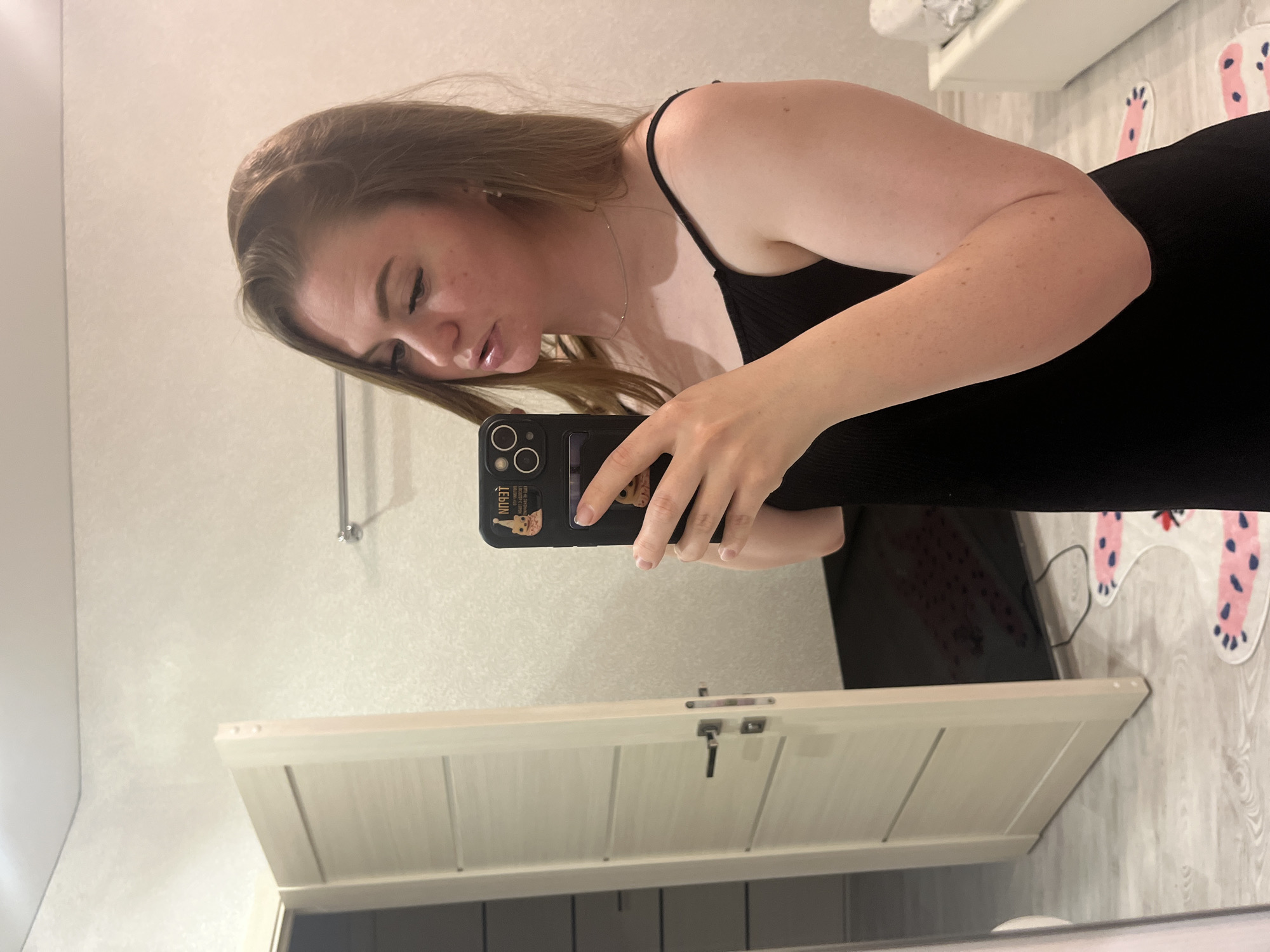 A woman is taking a selfie in a bathroom. She is wearing a black tank top. The background includes a white wall with a towel rack, a white door, a black object on the floor, and a white rug with pink paw prints.