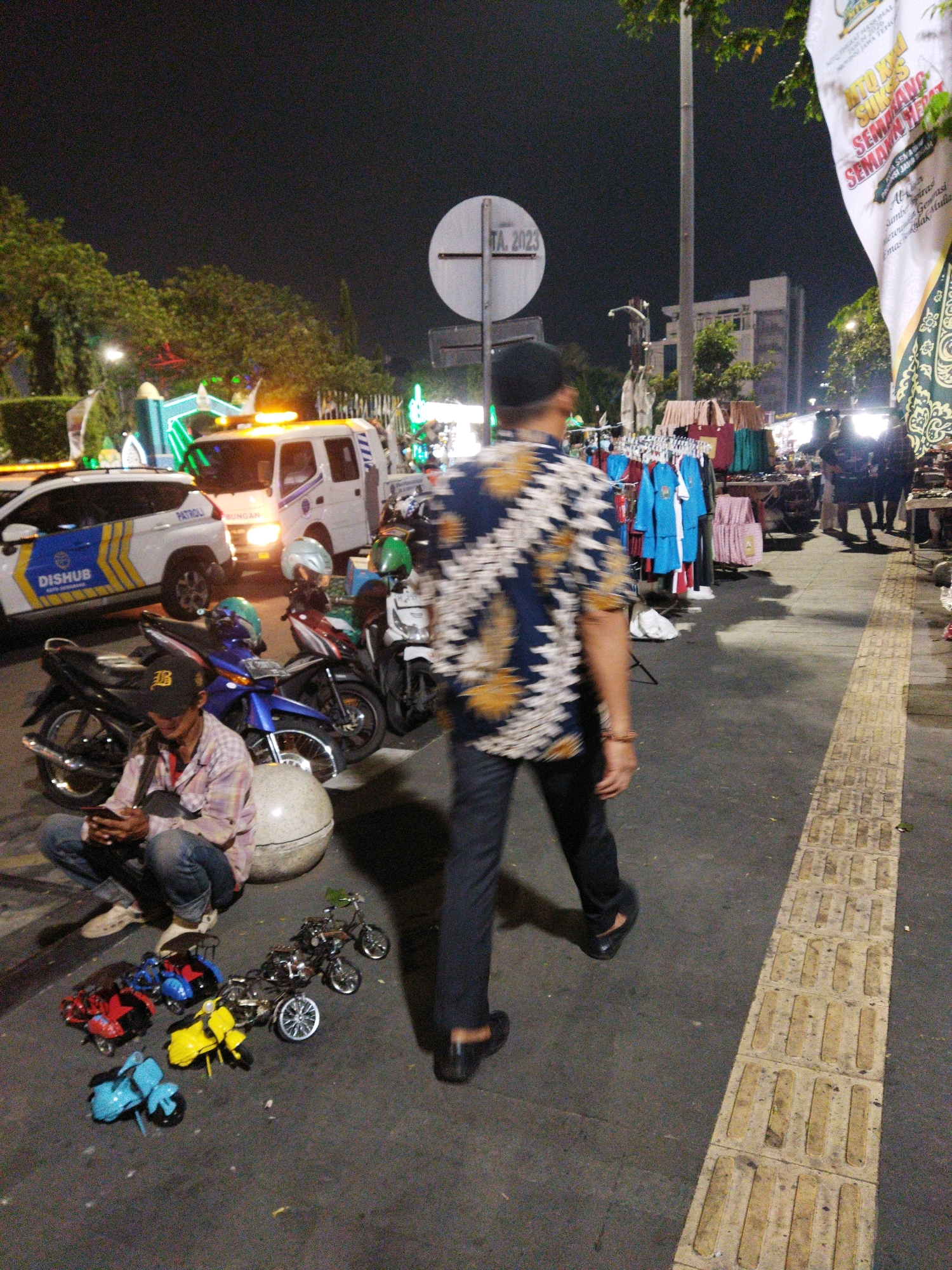 This nighttime street scene in Semarang, Indonesia, captures a vibrant roadside market. A person in a patterned shirt and black pants walks away from the camera, his form blurred by motion. In the foreground, a man wearing a plaid shirt, jeans, and a baseball cap is seated on the ground, looking at his phone. He is positioned near a large, spherical object and a collection of miniature toy motorcycles in various colors, including red, blue, yellow, and black, arranged on the asphalt.
Behind the seated man, several real motorcycles are parked, some with helmets placed on them. One motorcycle is blue, another dark-colored, and a third appears to be white. A white utility vehicle with flashing orange lights, possibly a tow truck or service vehicle, is visible to the right of a white patrol car with "DISHUB" (Transportation Agency) written on its side. The patrol car has blue and yellow stripes.
Further back, a market stall displays clothing on racks, with various garments, including what appear to be blue uniforms and pink items, hanging for sale. The market is illuminated by artificial lights, casting shadows on the ground. The environment suggests a busy urban setting at night, likely a popular gathering spot or a street with commercial activity. Trees line the background, and a sign with "TA. 2023" is visible on a pole. A large banner with text and decorative patterns is also present, hanging on the right side of the frame. The ground is paved with asphalt, and a tactile paving strip runs along the right edge of the walkway.