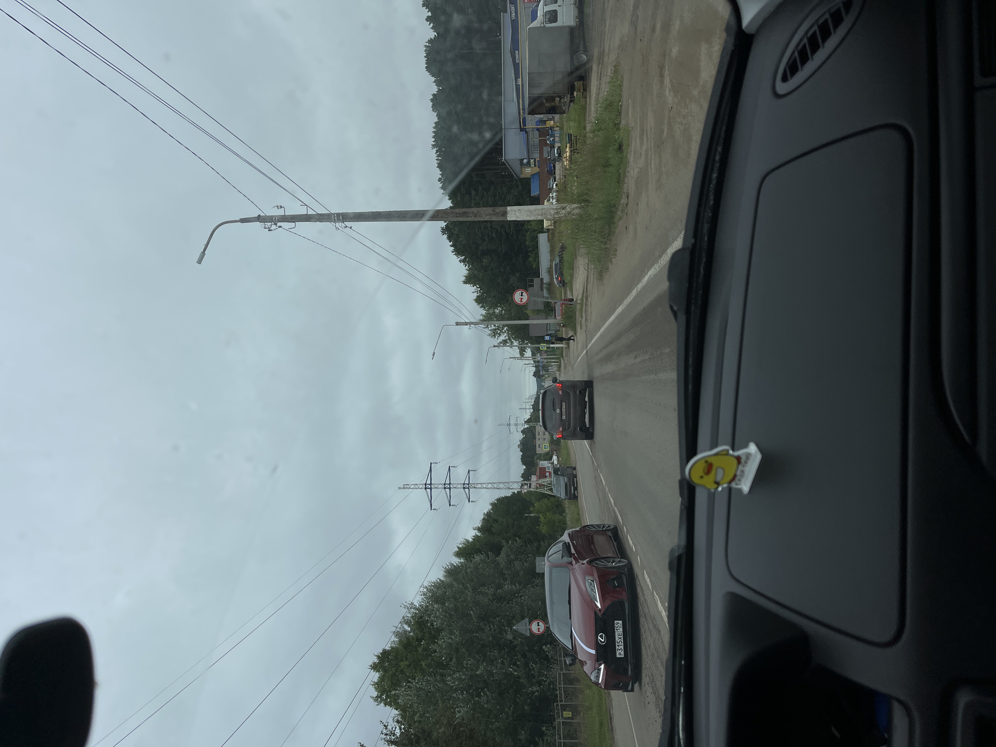 The image, captured from inside a vehicle, depicts a two-lane road in Chaikovsky, Russia. The sky is overcast with scattered clouds. On the left side of the road, a dark red Lexus sedan with a white stripe down its hood and the license plate P315XE 159 is in the foreground, traveling in the same direction as the camera. Further ahead on the same lane, a dark SUV is visible. On the right side of the road, a light-colored panel van is parked, with a large, flatbed truck positioned behind it. Adjacent to the road on the right, a forested area with tall trees lines the horizon, and a row of commercial buildings with various signage is present. Power lines are strung across the sky, supported by utility poles. A small, yellow duck-shaped air freshener is affixed to the dashboard of the vehicle from which the photo is taken.