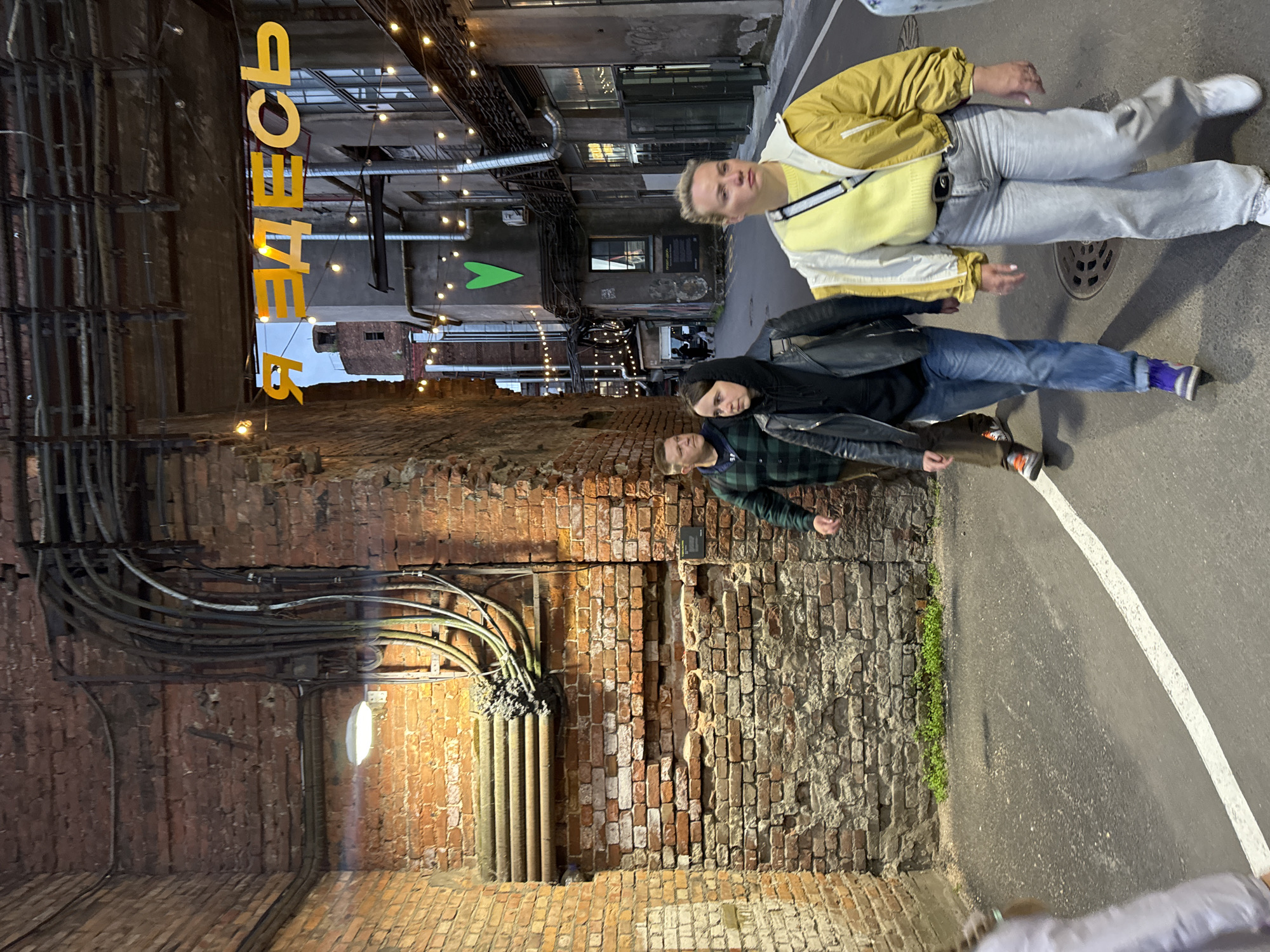 In Saint Petersburg, Russia, an outdoor scene unfolds in a narrow, industrial-style alleyway, likely part of a converted factory or warehouse district, featuring exposed brick walls and overhead industrial piping. A yellow neon sign, reading "Я ЗДЕСЬ" (which translates to "I'm Here"), hangs prominently above the walkway, illuminated by a string of small, bare lightbulbs that also adorn the buildings.
Three individuals are captured mid-stride, walking away from the camera. In the foreground, a woman with blonde hair wears a yellow and white jacket over a yellow top and light-wash jeans. To her left and slightly behind her, a person in a black leather jacket, black hoodie, and blue jeans walks with their head bowed. Further back and to the left, a man in a green plaid shirt and brown pants walks with his head turned to the side. A fourth person, partially visible on the far left, is seen from the back, wearing a white top.
The ground is paved with asphalt, marked by a white line that curves to the right. The lighting suggests it is dusk or evening, with the artificial lights creating a slightly moody ambiance. A bright green heart shape is painted on a grey building wall in the background, adding a touch of color to the otherwise muted tones of the architecture. The overall impression is one of urban exploration in a revitalized industrial space.