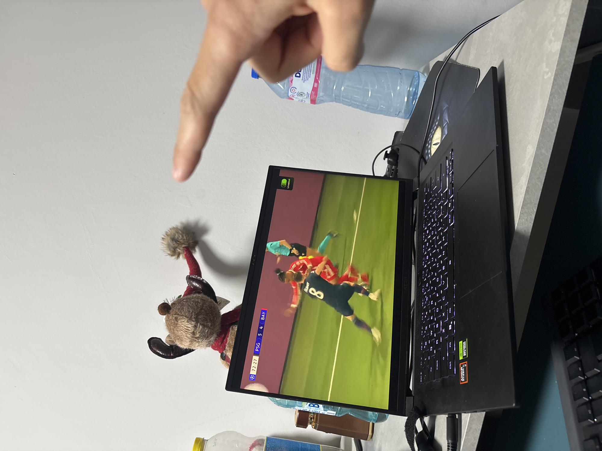 An indoor scene, likely in Plovdiv, Bulgaria, captures a person's hand pointing at a laptop screen displaying a live soccer match.
The central subject is a black laptop, open and positioned on a light grey, possibly concrete, desk or table surface. The laptop screen shows a soccer game in progress, with players in red and dark blue uniforms on a green field. Visible text on the screen includes "LiveBall" in the top left, the match time "72:27", and the score "PSG 5" vs "BAY 4". A player wearing jersey number "8" and named "HALMAY" is also discernible. The laptop's keyboard is illuminated with purple backlighting. On the laptop's body, "NVIDIA GeForce RTX" and "AMD Ryzen 9" logos are clearly visible.
To the left of the laptop screen, a small, furry, brown stuffed animal, resembling a reindeer and wearing a red knitted hat and scarf, is perched. Behind the laptop, a clear plastic water bottle with a blue cap and a brand label (implied to be "Devin" given the location context) is situated. A person's right hand is prominent in the foreground, pointing downwards towards the laptop screen. Various cables and a dark mousepad are also visible around the laptop. The scene suggests a casual setting for watching a sports event.