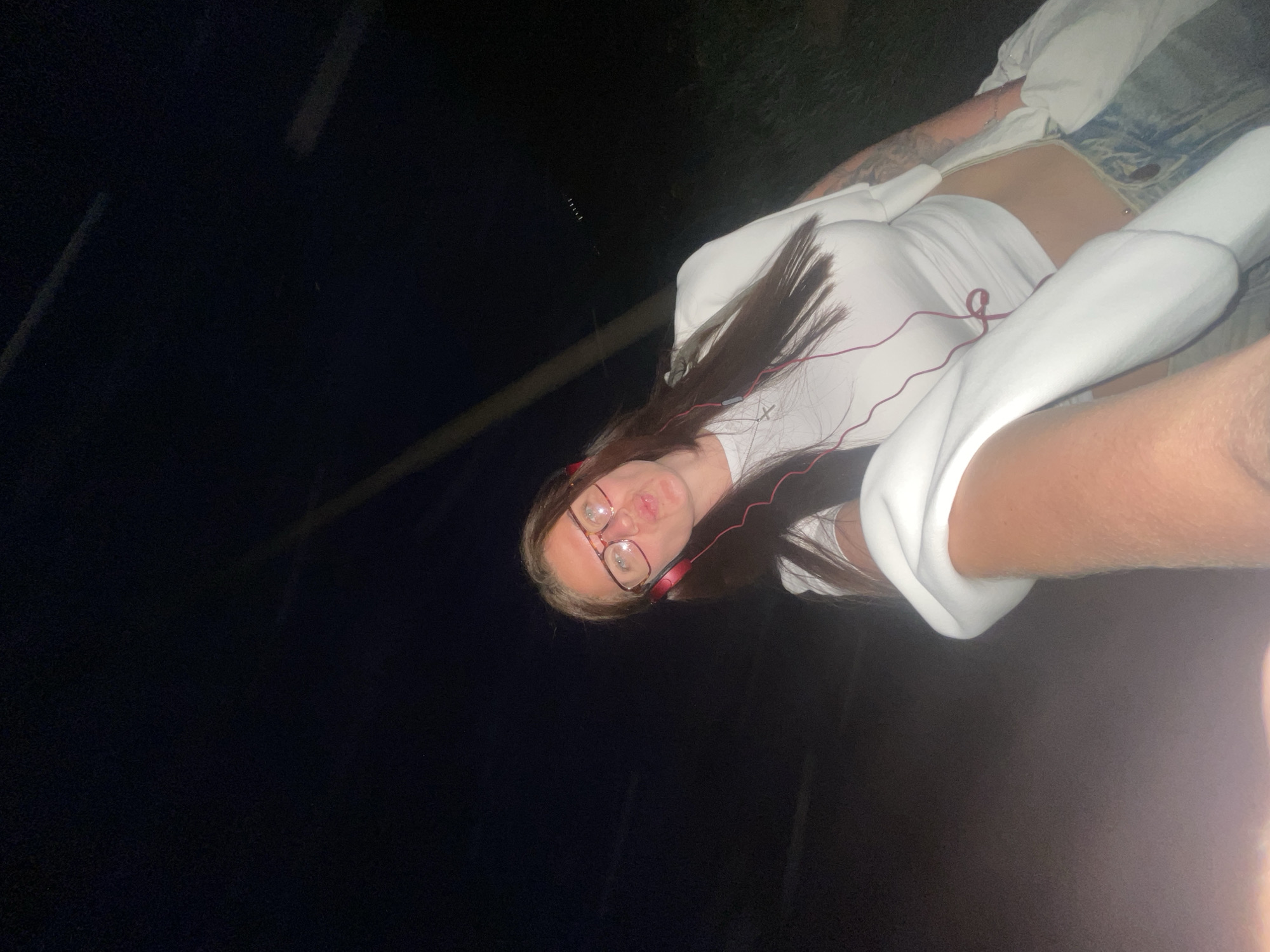 A young woman is posing for a photo outdoors at night. She is wearing glasses, headphones, a white t-shirt, and light-wash denim shorts. She has long, dark hair with blonde streaks in the front. Part of another person's arm is visible on the right side of the frame. The location is assumed to be in the city of Molokovo, Russia.