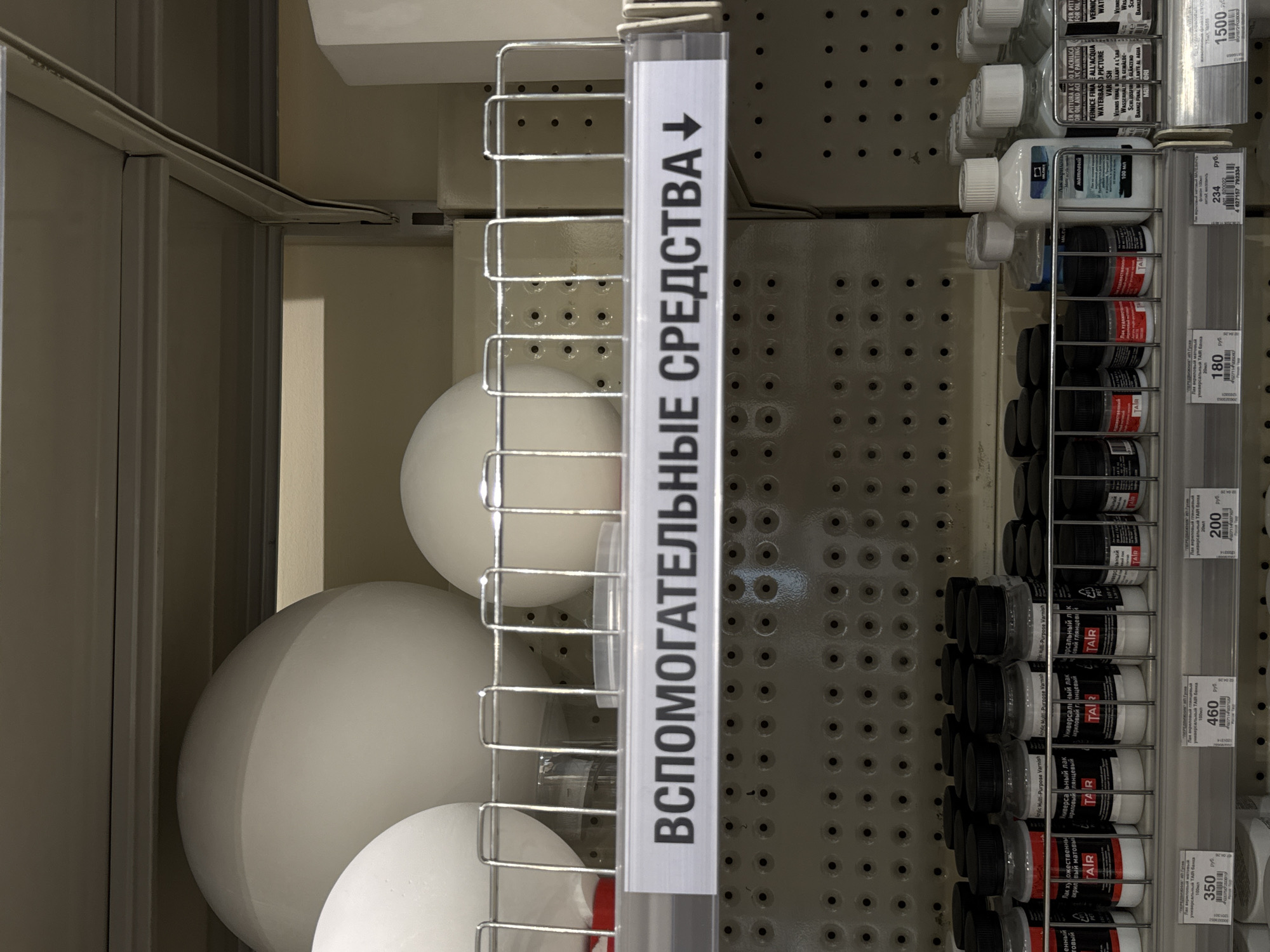 This is a vertical, eye-level shot of a retail store aisle, likely in Moscow, Russia. The focus is on a display rack featuring art supplies. The rack is made of silver metal wire and holds various small bottles and jars, most with black caps. Some bottles are white, and others appear to be clear glass. A prominent white label with black Cyrillic text is attached to the rack, reading "ВСПОМОГАТЕЛЬНЫЕ СРЕДСТВА↓" which translates to "AUXILIARY MEANS" with a downward arrow, indicating the category of products.
Below the labels on the rack, price tags are visible. One tag shows "1500", another "234 руб.", "460 руб.", "200 руб.", "180 руб.", and "350". The text on some of the product labels is also partially legible, mentioning "TAIR" and various types of varnish and paint, such as "Универсальный лак акриловый глянцевый" (Universal acrylic varnish glossy) and "Лак художественный акрил вый матовый" (Artistic acrylic varnish matte).
In the background, shelves with more products are visible, along with a textured wall with small circular holes, characteristic of pegboards often used in retail displays. To the left of the main display rack, large, matte white spheres are partially visible, adding an unusual element to the scene. The lighting is bright and even, suggesting an indoor retail environment. No people are visible in the image. The overall impression is that of a well-organized retail display within an art supply store.
