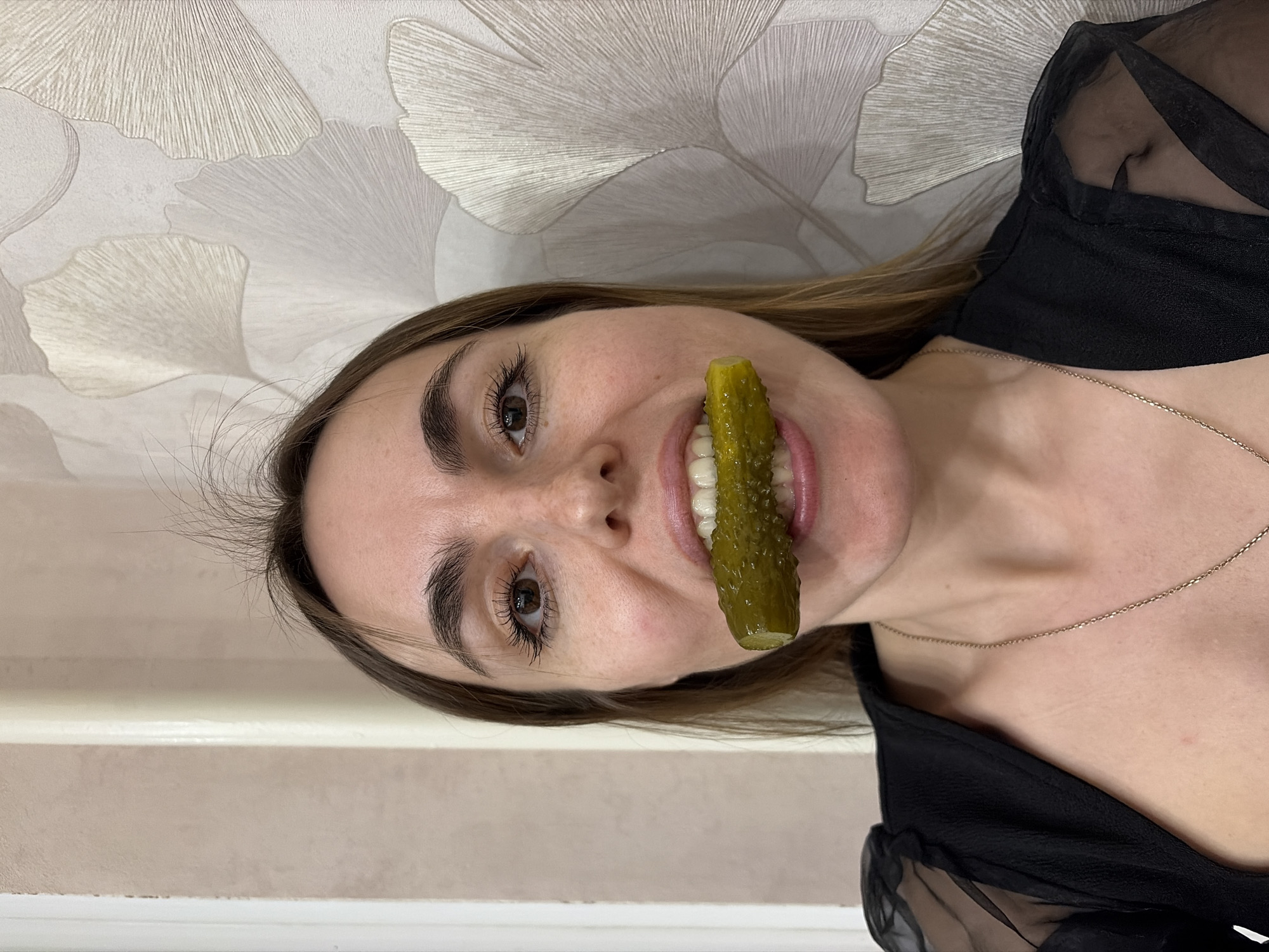 A woman is pictured indoors with a pickle held in her mouth. She has light skin, brown hair, and brown eyes. She wears a black top and a delicate gold chain necklace. The background includes a wall with a light-colored, patterned wallpaper and a portion of a cream-colored wall or surface. The image is likely taken indoors, but there is no way to determine if this is in Izhevsk, Russia without further information.