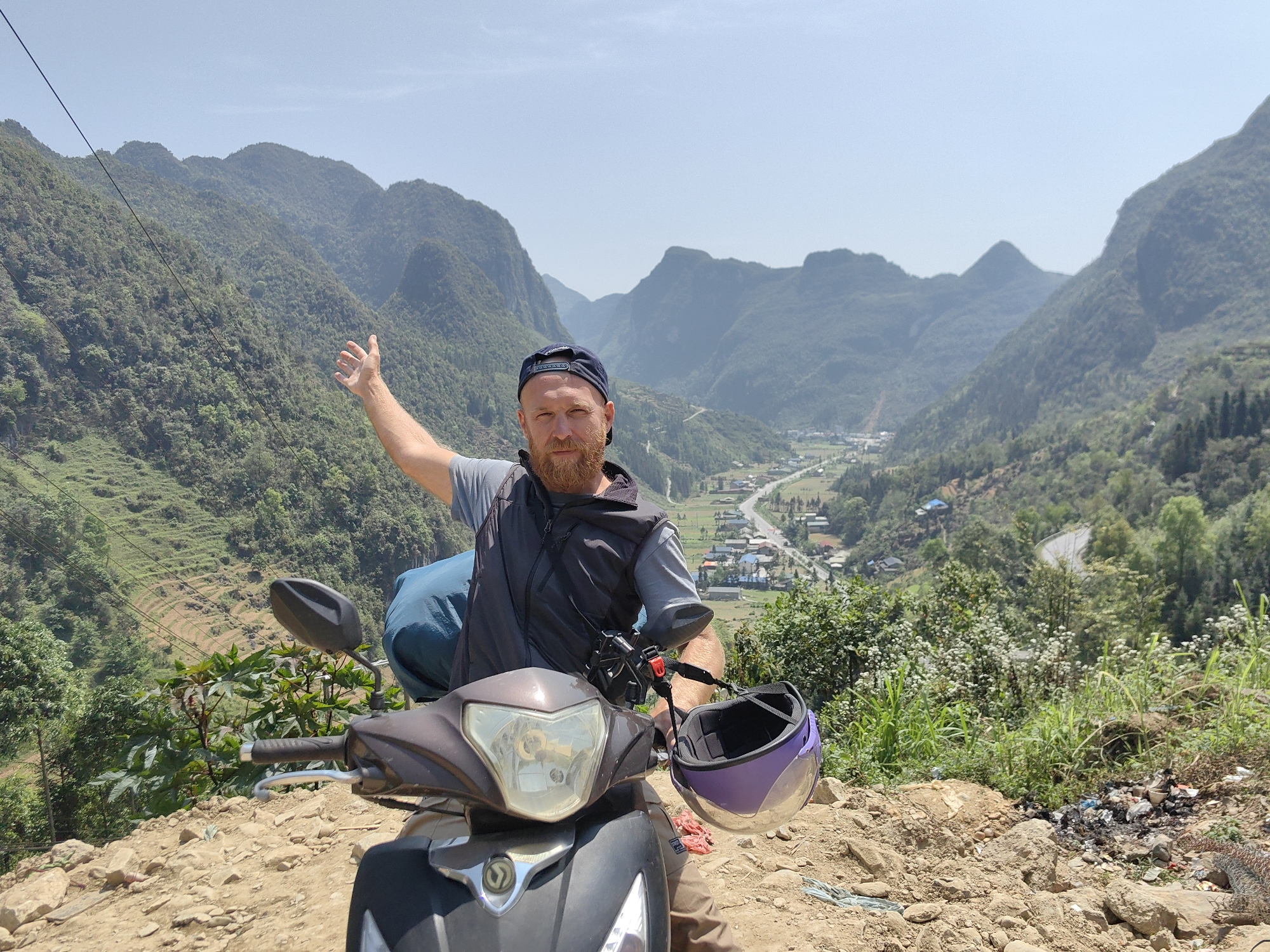 A man is sitting on a scooter parked on a rocky hill overlooking a valley in Phố Bảng Commune, Vietnam. The man has a beard, a cap, and is wearing a grey t-shirt and a dark vest. He is raising his left arm towards the hills. A purple helmet is hanging from the scooter's handlebar. In the background, there are mountains covered in trees and a valley with a town and a road running through it. In the foreground, there is vegetation and some trash. The sky is clear.