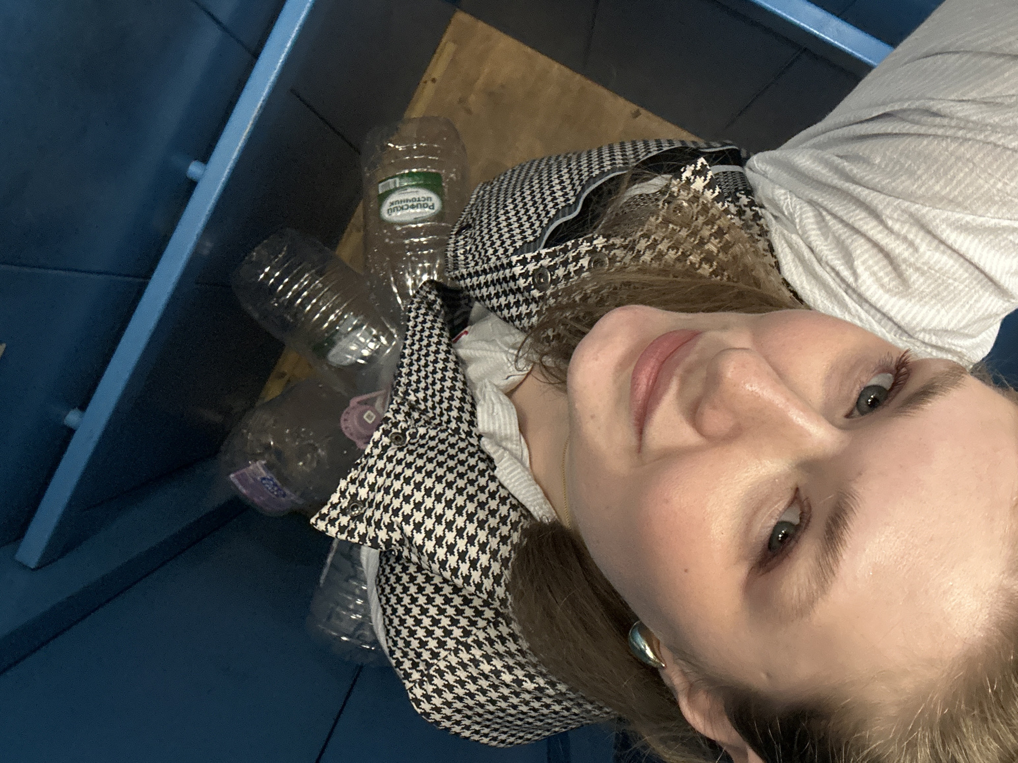 The image shows a woman in Kazan, Russia, looking into a blue recycling bin filled with empty plastic bottles. The bin's interior is visible, revealing a mix of clear and slightly tinted plastic bottles. The woman is wearing a houndstooth jacket over a light-colored shirt and a silver earring. The floor of the location appears to be made of wood.