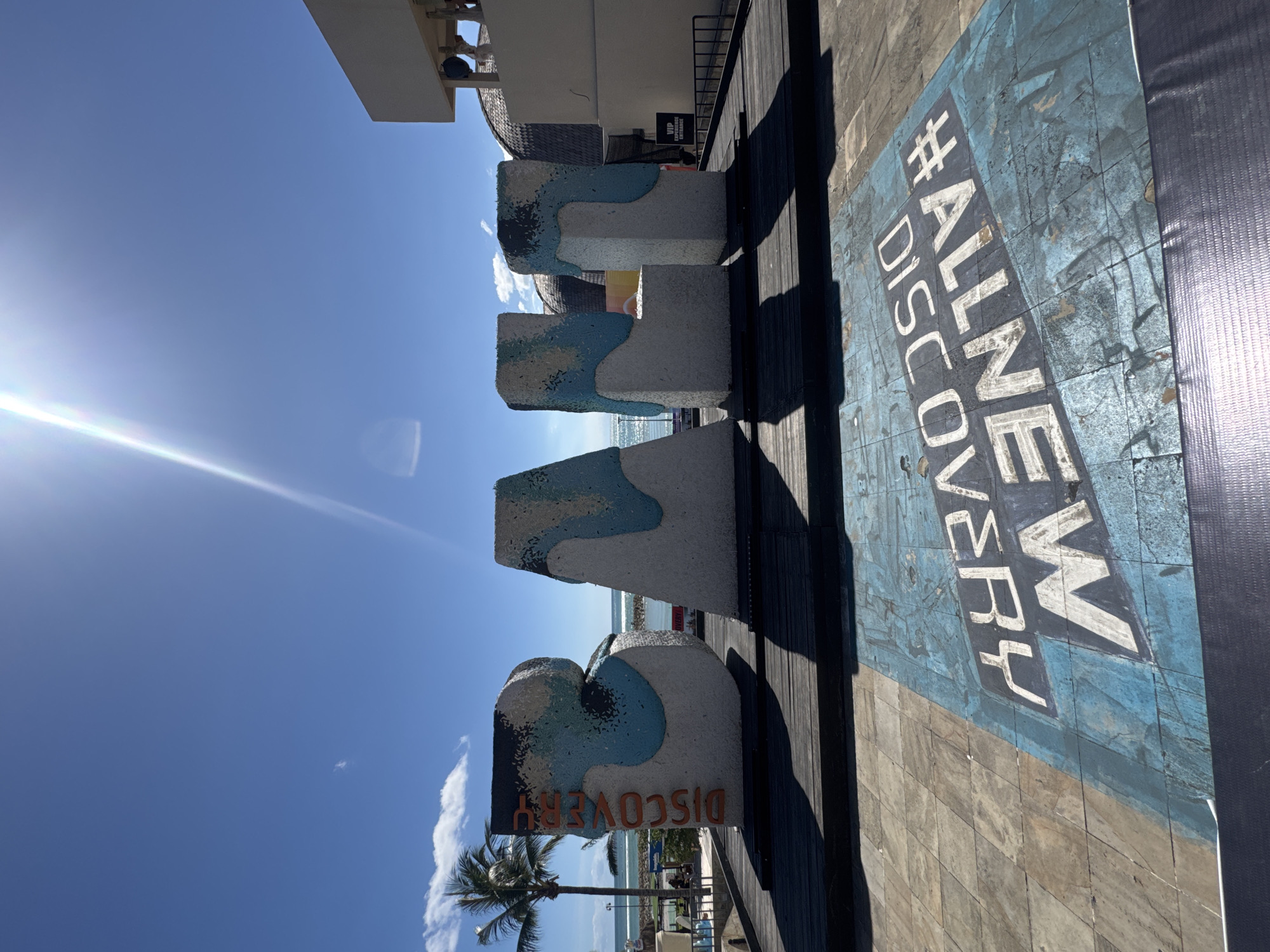 The image shows a sunny outdoor scene in Kuta, Indonesia, indicated by the large "DISCOVERY" sign made of concrete letters, with a design of blue and white suggesting water. The letters stand on a wooden deck. Next to the letters is a painted sign on the deck with the text "#ALLNEW DISCOVERY". In the background, there is a palm tree, the ocean, and some buildings. The scene suggests a tourist area or a landmark within Discovery Shopping Mall in Kuta. The sun is bright, casting a lens flare in the sky.