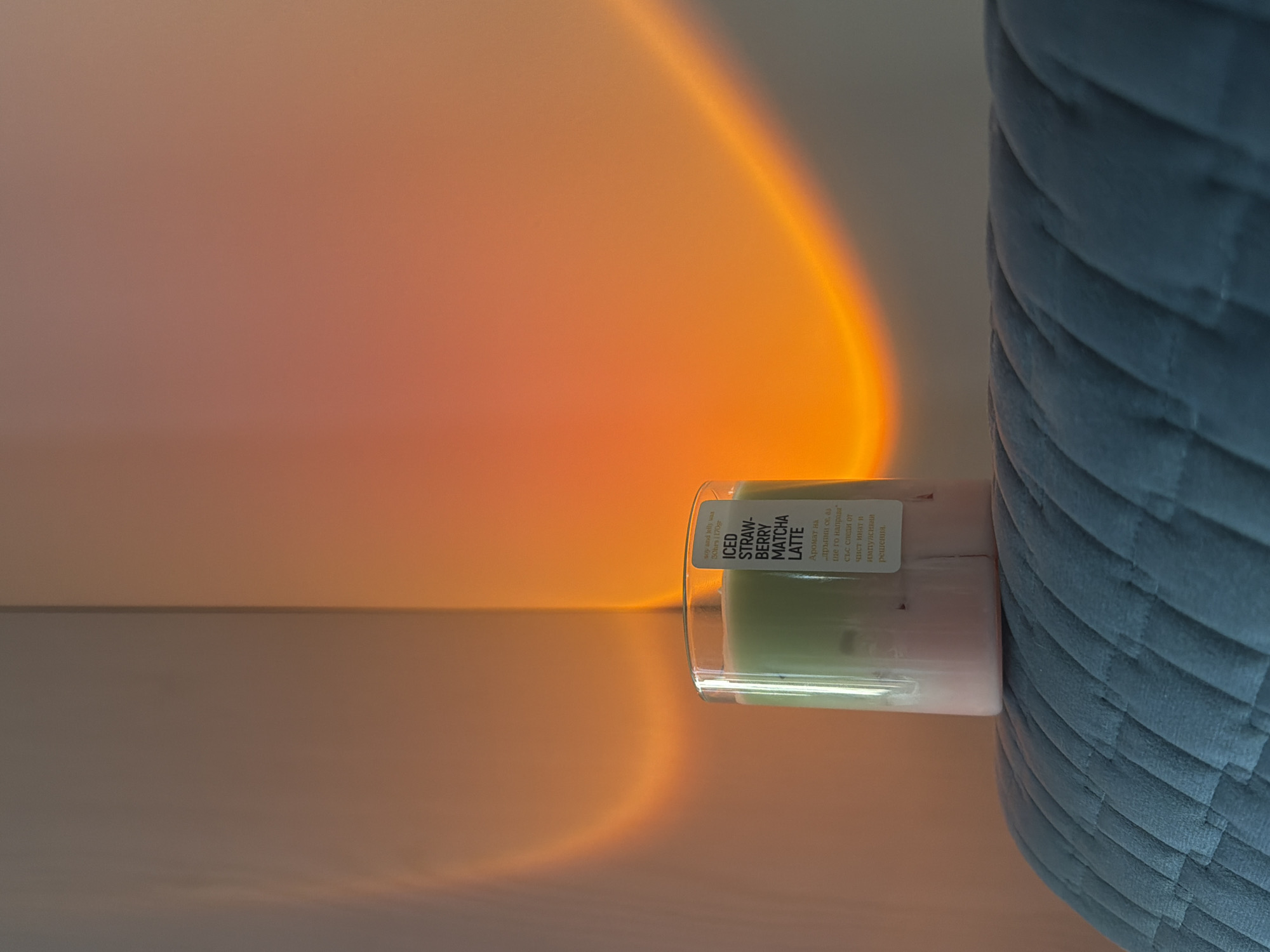 Here is a description of the image:
The image shows a candle with an "Iced Strawberry Matcha Latte" label next to a blue quilted chair. The background is a gradient of orange, giving the impression of a sunset-like effect. The text on the candle is in both English and Bulgarian. The candle is placed on what appears to be a flat surface, possibly a table or floor.