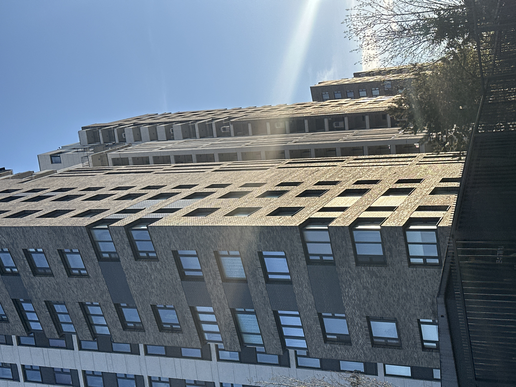 The image shows a multi-story building in Moscow, Russia, taken from a low-angle perspective. The building has a predominantly gray brick facade with regularly spaced windows. The windows reflect the blue sky. The building’s design incorporates horizontal ledges or balconies. Above the brick facade is a section with lighter colored concrete or stone. The sky is a clear blue with some glare from the sun at the top. In the upper right corner is a tree branch and a fire escape ladder.