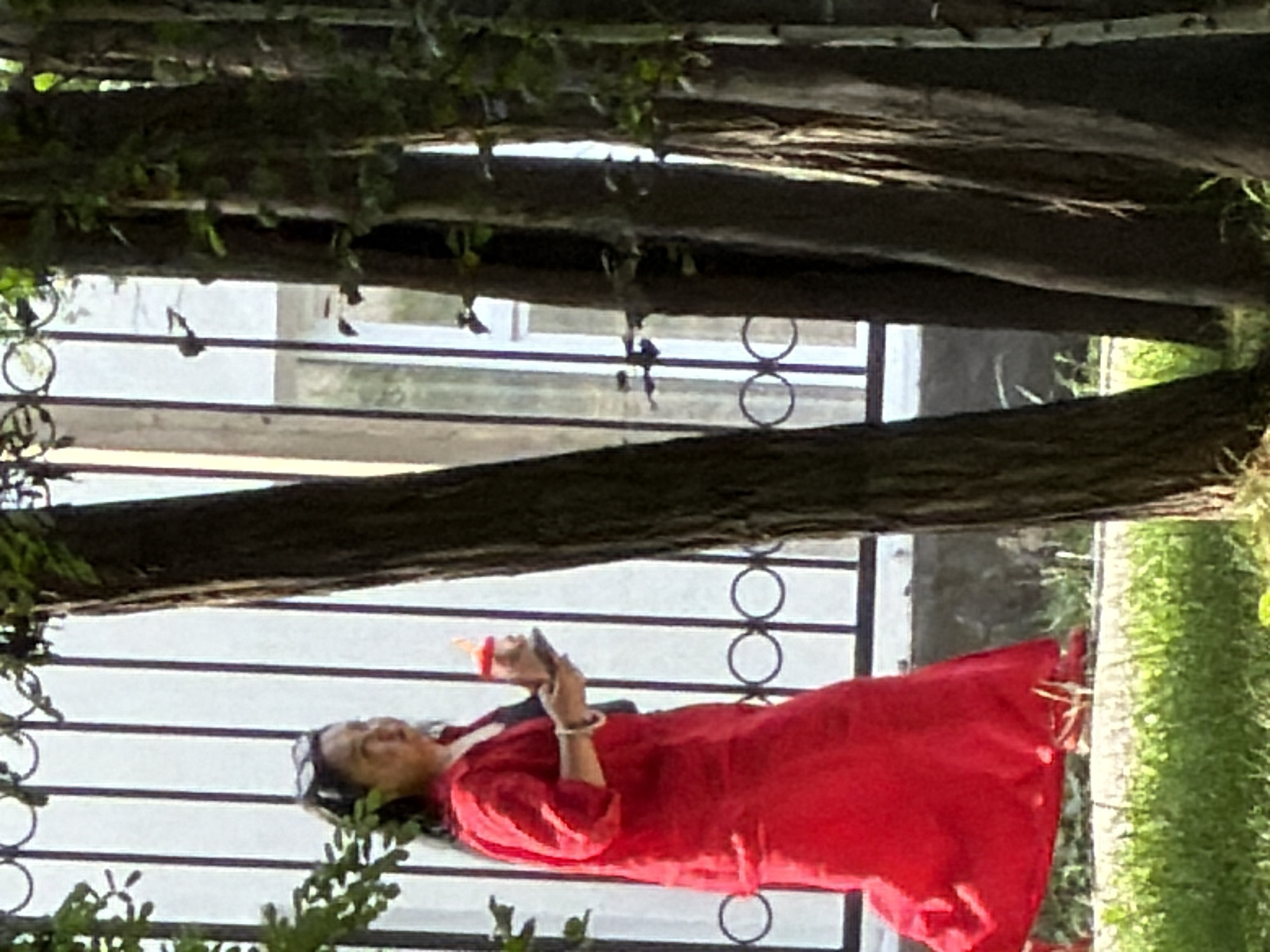 A woman in a bright red dress walks outdoors next to a black metal fence with circular accents. She holds a dark-colored phone in her right hand and a small orange bottle with a white cap in her left hand. She wears sunglasses on top of her head and a bracelet on her left wrist. The background shows a white building with vertical window panes and a black fence. Trees with green leaves are in the foreground, obscuring part of the scene. The ground is covered with green grass.