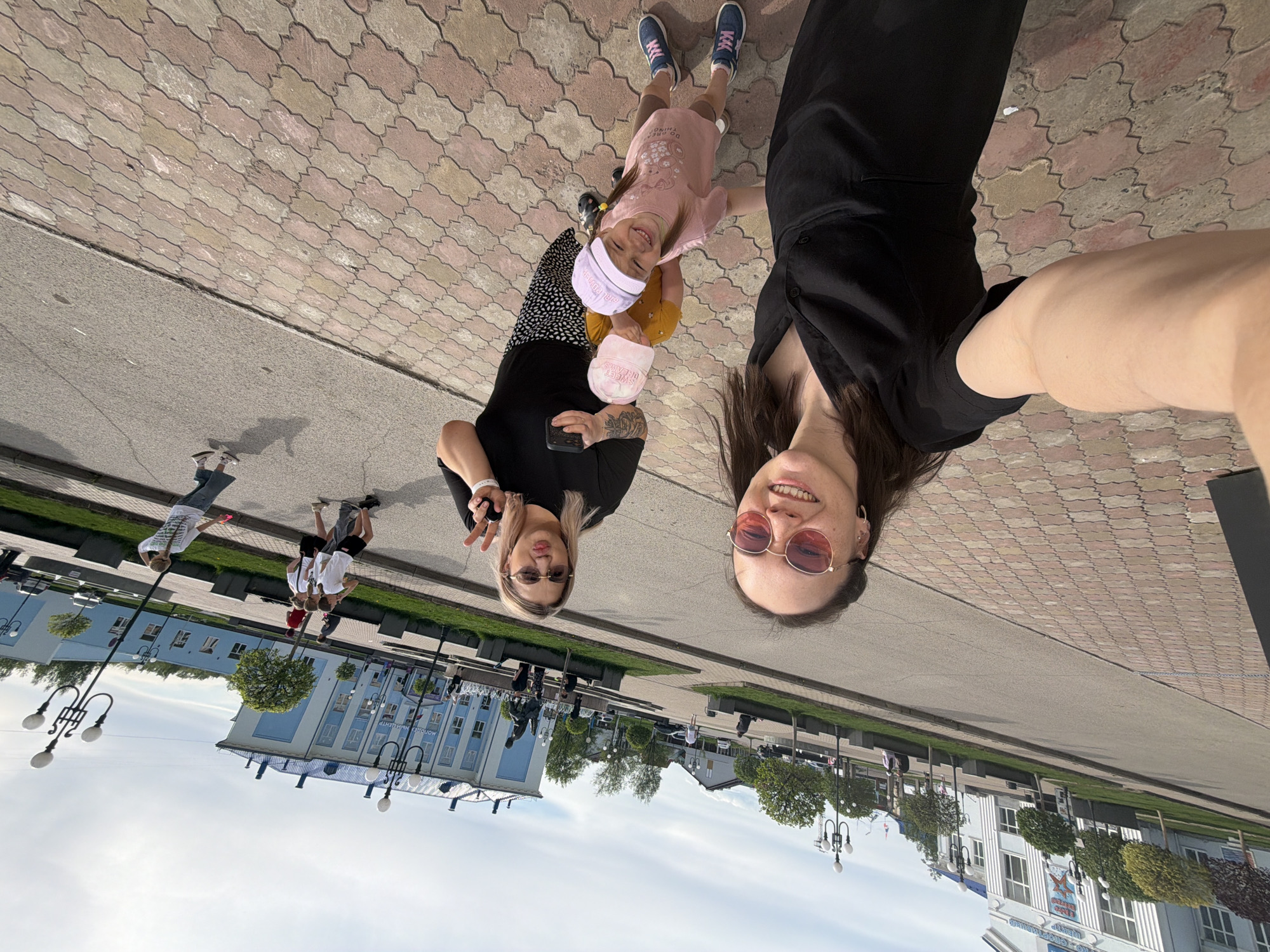 The image is a selfie of two women and a young girl on a brick-paved area near a street in Chaikovsky, Russia. The woman on the right is holding the camera, wearing round sunglasses and a black top. The other woman, to the left, is making a peace sign, wearing rectangular sunglasses and a black and white polka dot dress. The young girl is between them, wearing a pink dress and two hats. In the background, there is a large building with blue and white exterior. People are walking along the street. The sky is cloudy. Lampposts are present. The location is identifiable as Chaikovsky, Russia due to the building with Cyrillic script.