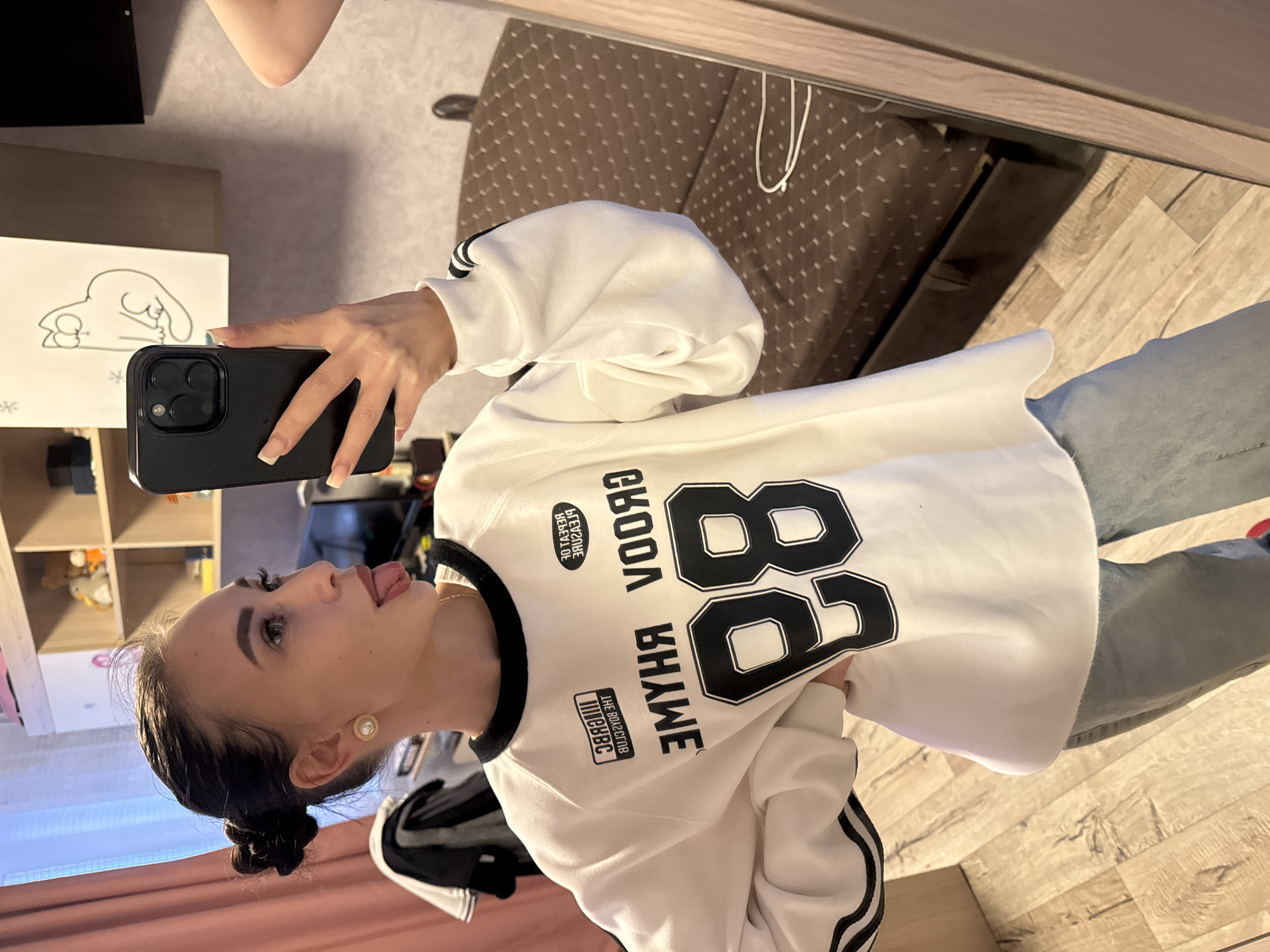 A young woman is taking a selfie in what appears to be a bedroom. She is wearing a white sweatshirt with black lettering and the number 88, paired with blue jeans. The room contains a bed with a patterned bedspread, a shelf with various items, and a window. The location is indicated as the city of Chaikovsky, Russia.