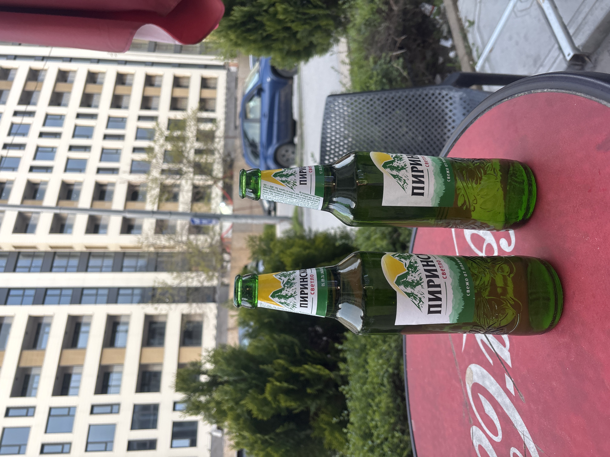 The scene depicts two bottles of "Pirinsco" beer on a red table top with a partially visible "Coca-Cola" logo. The bottles are green and labeled in Cyrillic. Behind the table, there is foliage, a building with a grid-like facade, and a blue car. The location is likely a cafe or outdoor setting in a urban environment. A portion of a red awning is visible at the top left corner of the image. Based on the "Pirinsco" beer bottles, the image is likely from Bulgaria. Without further information it cannot be stated with certainty that the location is Plovdiv.