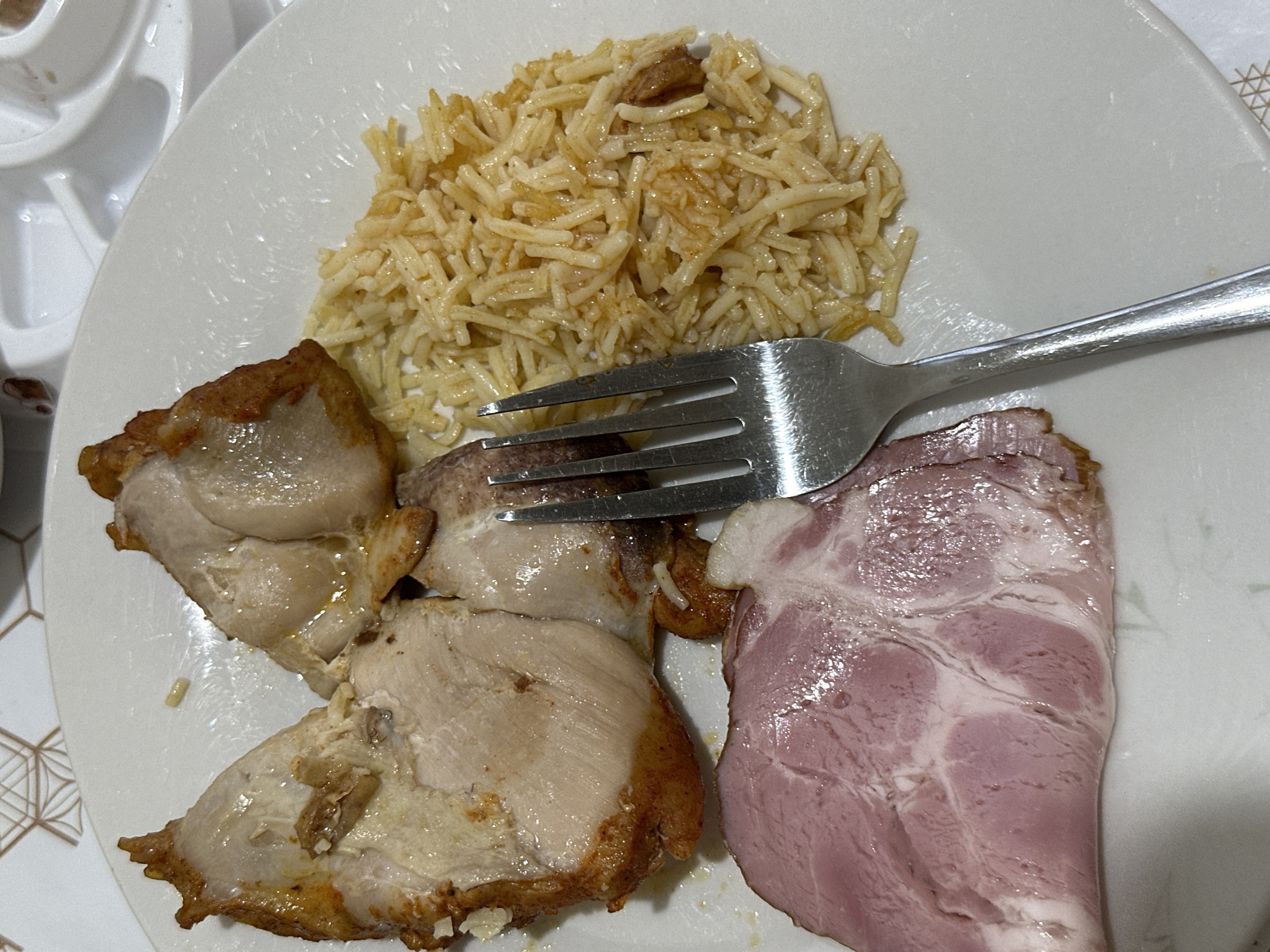 The image shows a plate of food, likely a meal. The plate contains a portion of rice, several pieces of cooked meat, and a slice of ham. A fork rests on the plate, its tines partially embedded in the rice. The food appears to be served on a white, circular plate, which is placed on a surface with a geometric pattern, possibly a tablecloth or placemat. The lighting suggests an indoor setting, potentially a dining room or kitchen, and the time of day is not discernible. There is no visible text in the image. The overall impression is of a casual meal, with the food presented in a straightforward manner. The context provided, Budva, Montenegro, suggests this meal was consumed in that location, but there are no specific visual cues within the image to confirm this.