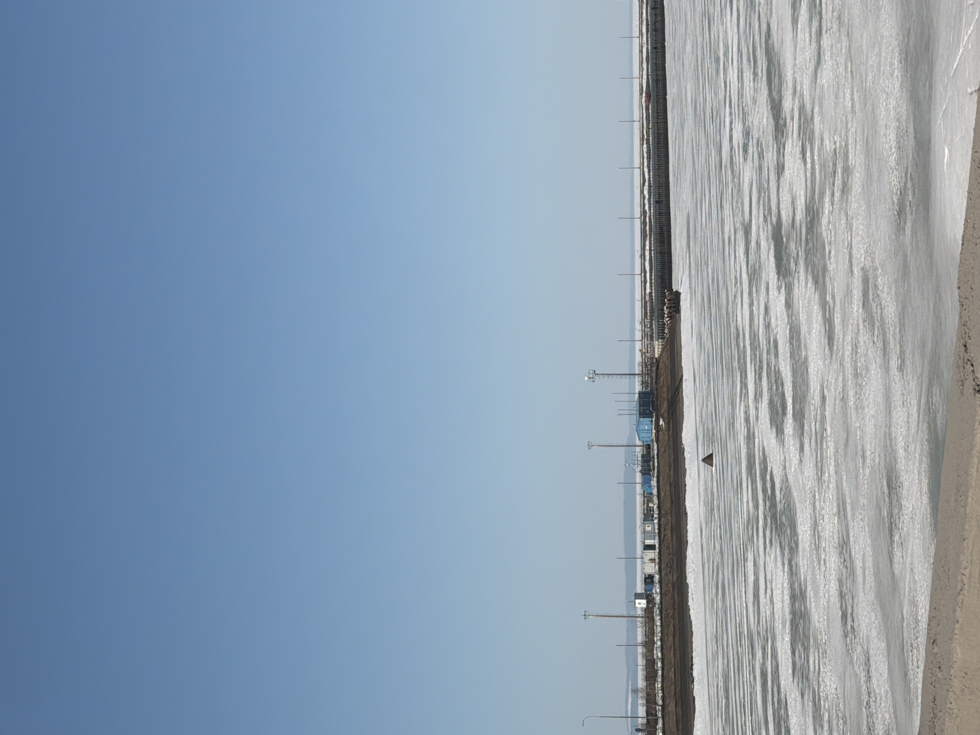 The image depicts a frozen body of water adjacent to a coastal area with infrastructure. The sky is clear blue.
Along the coast, there are utility poles, some structures appearing to be buildings or containers, and fencing. A dark brown dirt area separates the infrastructure from the frozen water.
The surface of the frozen water has a textured appearance, with patterns that suggest wind action on the ice. A single, dark triangular object sits in the middle of the ice.
It is impossible to confirm the specific location in the city of Chaikovsky, Russia, without further information.