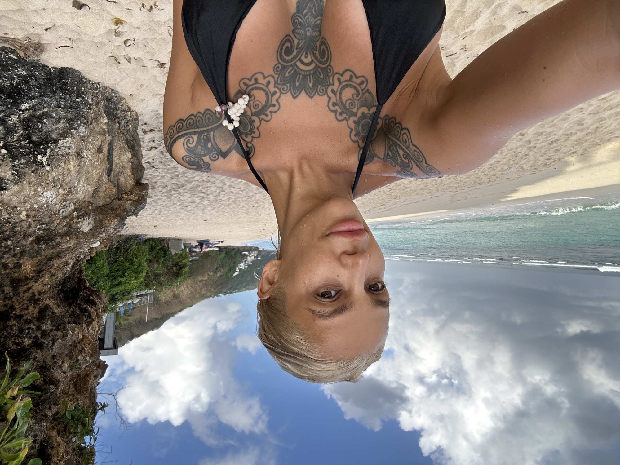 A woman with blonde hair and extensive tattoos on her chest and shoulders takes a selfie on a sandy beach. She is wearing a black bikini top adorned with white beads on the left strap. The background shows a turquoise ocean with waves, a sandy beach, a rocky cliff face on the right, and a vegetated hillside with buildings in the distance. The sky is partly cloudy with a bright blue sky visible. The location is Kuta Selatan, Indonesia.