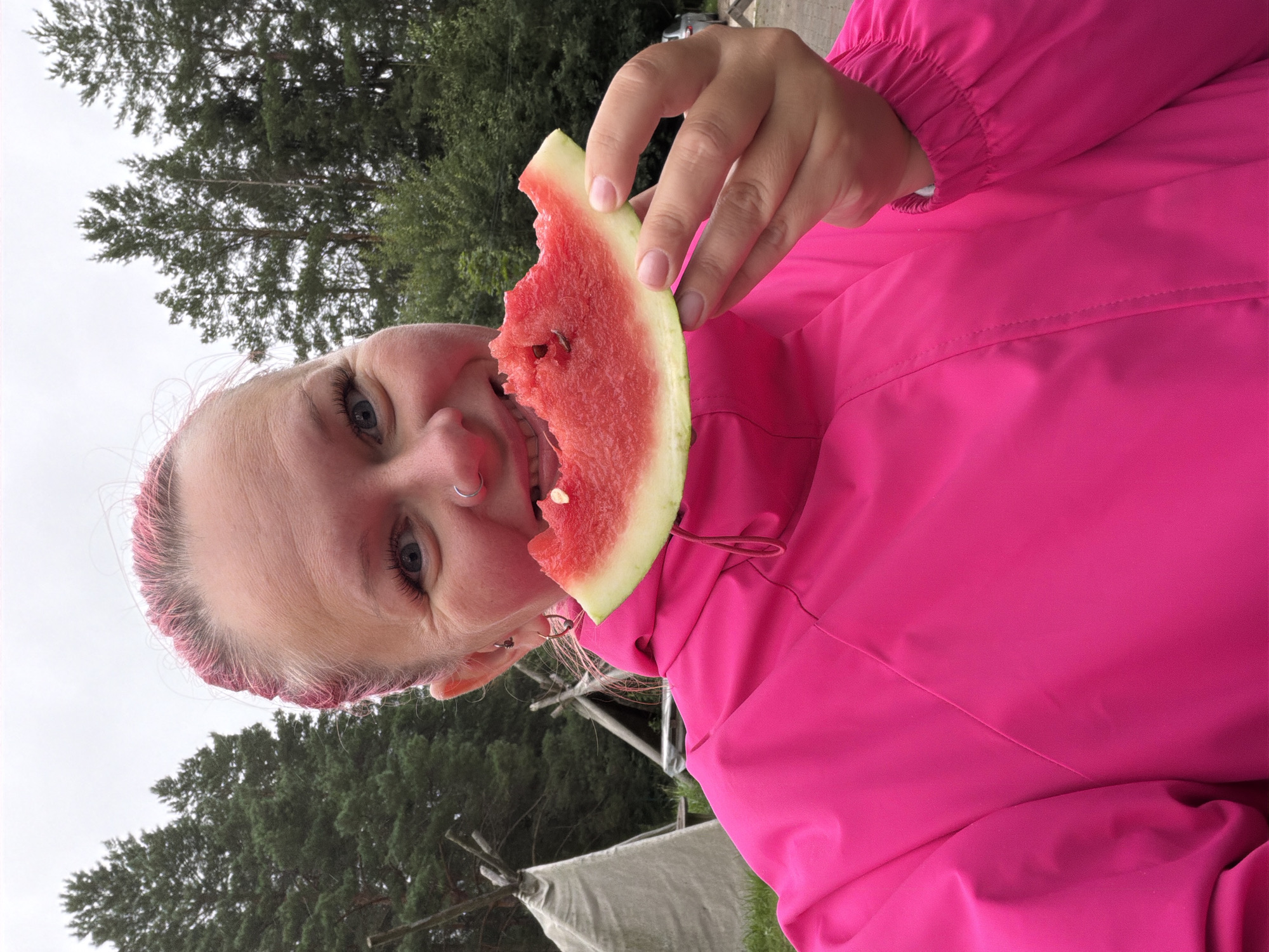 A woman with bright pink hair is taking a selfie while eating a slice of watermelon. She is wearing a bright pink jacket and has a nose ring and earrings. Behind her, there are trees and a teepee-like structure. The location appears to be an outdoor setting, possibly a campsite or park, in a natural environment with trees and a cloudy sky. The background suggests a rural or semi-rural area within or near Прикамский, Russia.
