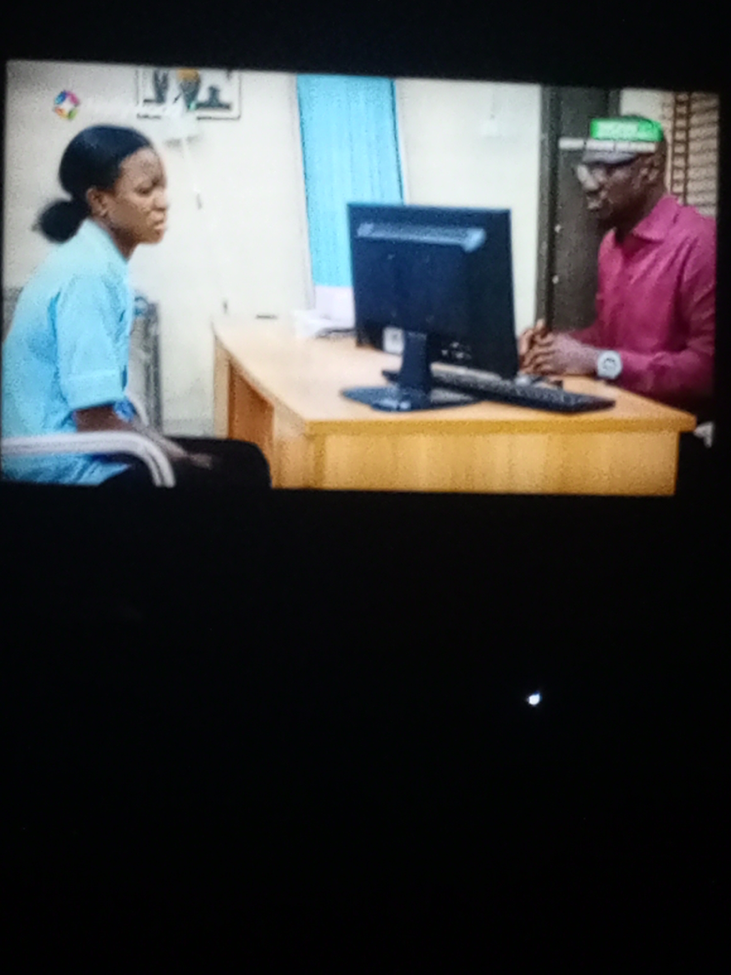 In an indoor setting, a woman in a light blue uniform is seated on the left, facing a man with dark skin wearing glasses and a pink shirt. He also wears a hat with green letters. They are positioned on opposite sides of a wooden desk with a computer monitor and keyboard on it. The location is Unknown, Unknown. The background features a window with blue curtains and a framed picture on the wall.