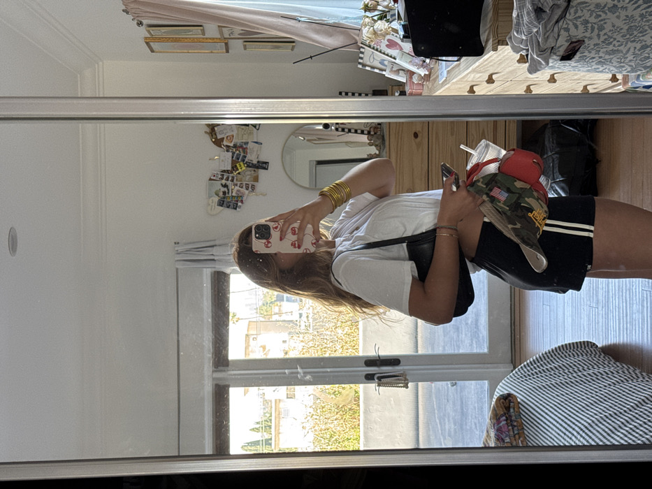 A female subject is depicted taking a mirror selfie indoors, facing left. She has long, wavy light brown hair and is dressed in a white short-sleeved t-shirt and black athletic shorts with white side stripes. A black shoulder bag hangs on her left side. Her right wrist displays multiple gold-toned bangles, while her left wrist has thinner bracelets. She holds a smartphone with a light pink, cherry-patterned case in her left hand, positioned for the photograph. Her right hand concurrently holds a camouflage baseball cap featuring an American flag patch, a red baseball cap, and a clear plastic cup with a lid and straw.
The indoor setting appears to be a residential room with white walls and a light-colored wooden floor, illuminated by natural light. To the subject's left, a bed with a striped white and dark green/blue duvet is partially visible. Beyond this, large double glass doors with dark frames and white vertical mullions reveal an outdoor scene. To the subject's right, a light-toned wooden dresser with multiple drawers is present, topped with framed pictures and various decorative items. A small circular mirror is affixed to the wall above the dresser, and a heart-shaped collage of images or notes is visible on the adjacent wall. Light pink curtains are partially visible on the far right.
Through the exterior glass doors, an urban or suburban landscape is visible under bright daylight. The scene includes buildings with stucco exteriors, lush green foliage, and distinct palm trees. This architectural style, vegetation, and sunny climate indicate a location consistent with Los Angeles, California, United States of America.