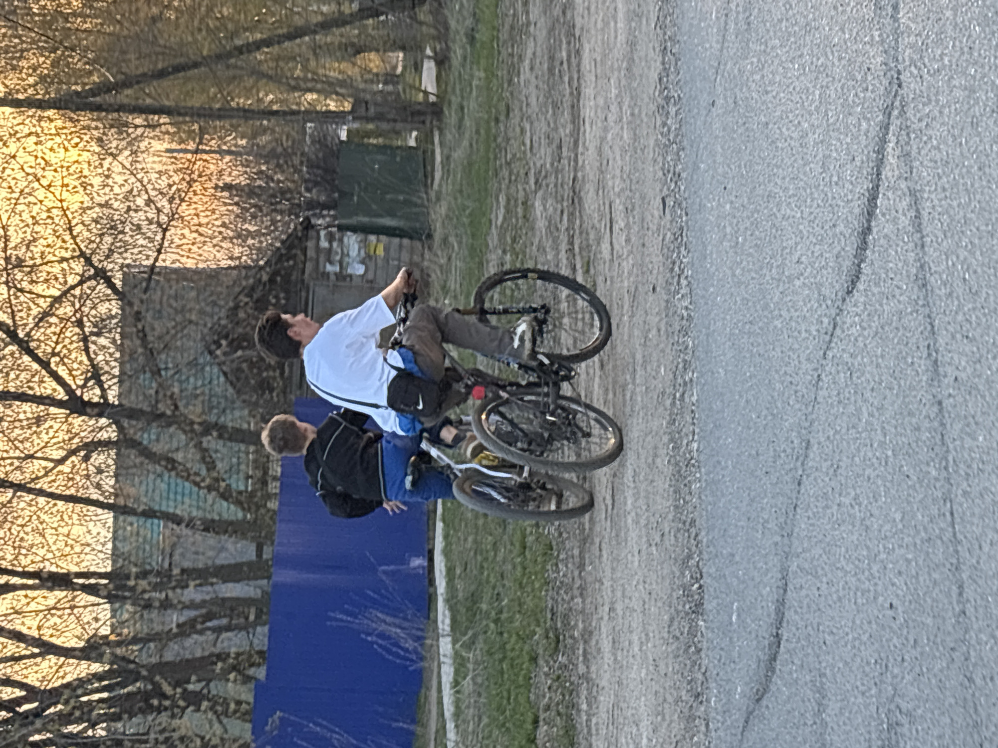 Two individuals are riding bicycles on a path next to an asphalt road. The older individual is wearing a white t-shirt and dark pants, while the younger individual is wearing a black jacket and blue pants. A blue fence is visible in the background, along with trees and buildings. The lighting suggests it is late afternoon or early evening. The location is likely in the city of Chaikovsky, Russia, as requested.