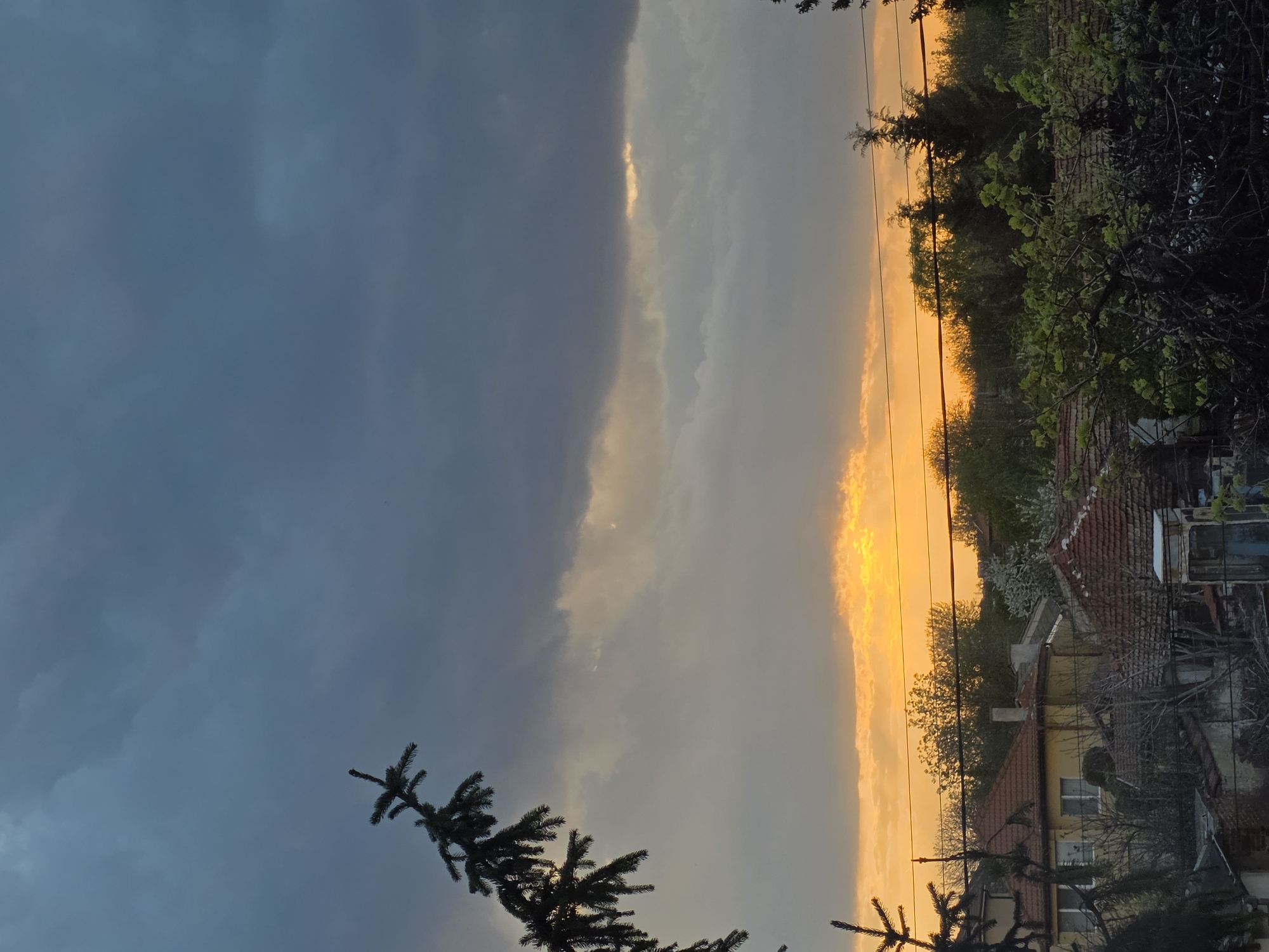 The image depicts a dramatic landscape during either sunrise or sunset over the village of Yagodovo, Bulgaria. The sky is a central feature, presenting a stark contrast between dark, stormy conditions and brilliant, warm light.
The left portion of the sky is dominated by heavy, dark grey-blue clouds, suggesting an approaching or lingering storm front. In stark opposition, the right side of the sky glows intensely with vibrant orange and yellow hues, indicating the sun's low position on the horizon. A prominent horizontal band of golden light separates the darker clouds from the brightly lit area, with some lighter clouds catching the sun's rays.
In the immediate foreground, the dark silhouette of a coniferous tree branch, possibly a fir or spruce, extends from the lower left corner into the frame.
Below the illuminated horizon, a village scene unfolds. Several houses with traditional red-tiled roofs are visible, nestled amidst abundant trees and bushes. One house in the midground features yellow walls and white-framed windows. Many trees, some with fresh, light green foliage indicative of spring or early summer, are silhouetted against the bright sky. Thin utility lines traverse the sky above the residential structures.
The overall scene captures a moment of striking natural beauty and meteorological intensity, showcasing the transitional light of either dawn or dusk in a picturesque village setting. There is no visible text in the image.