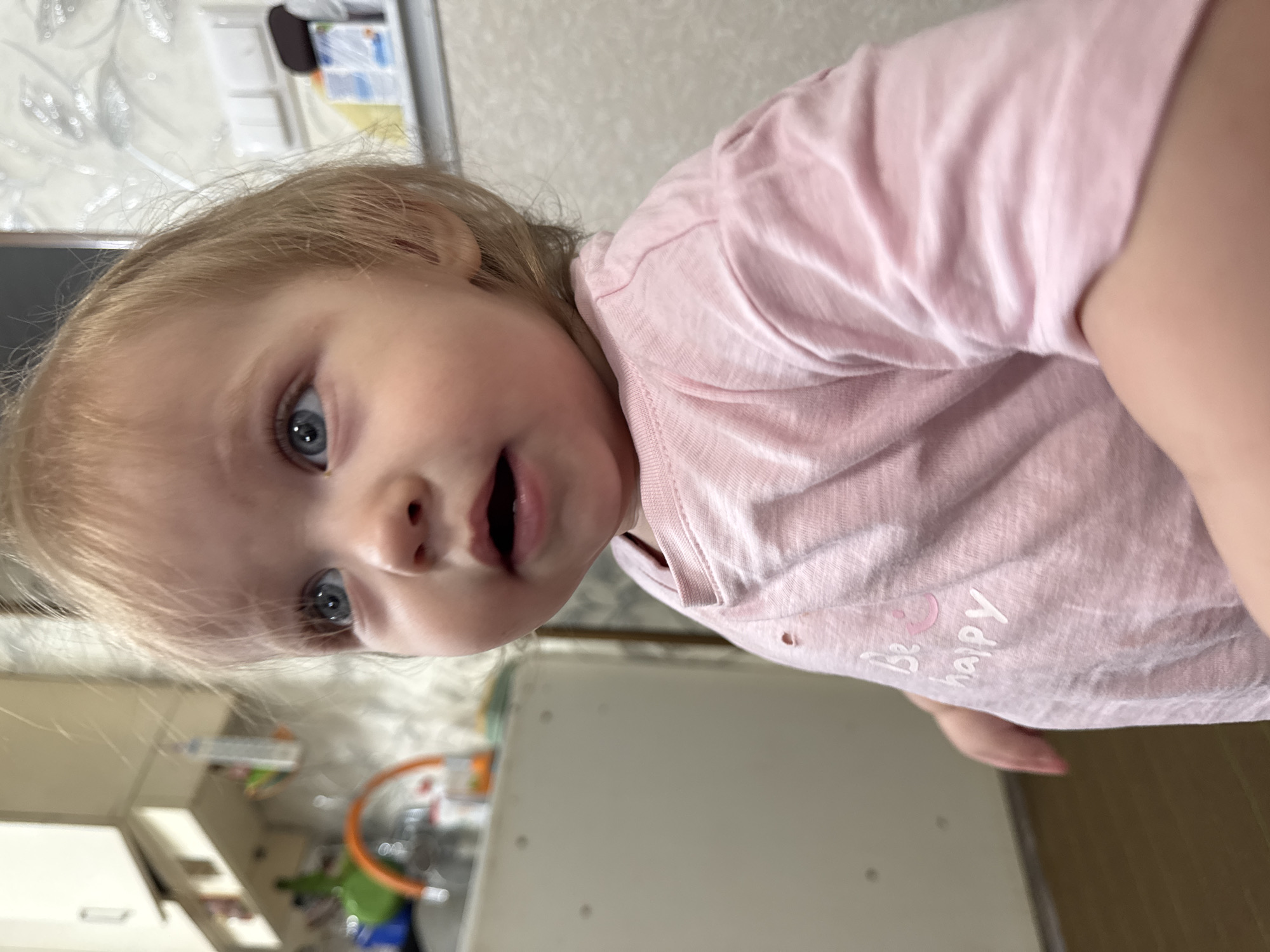 A young child with blonde hair and blue eyes wearing a pink t-shirt with the words "Be happy" on it is looking at the camera with an open mouth. The background is blurry but appears to be a kitchen with light-colored cabinets and a patterned wall. The image was taken in Nizhnyaya Gar', Russia.