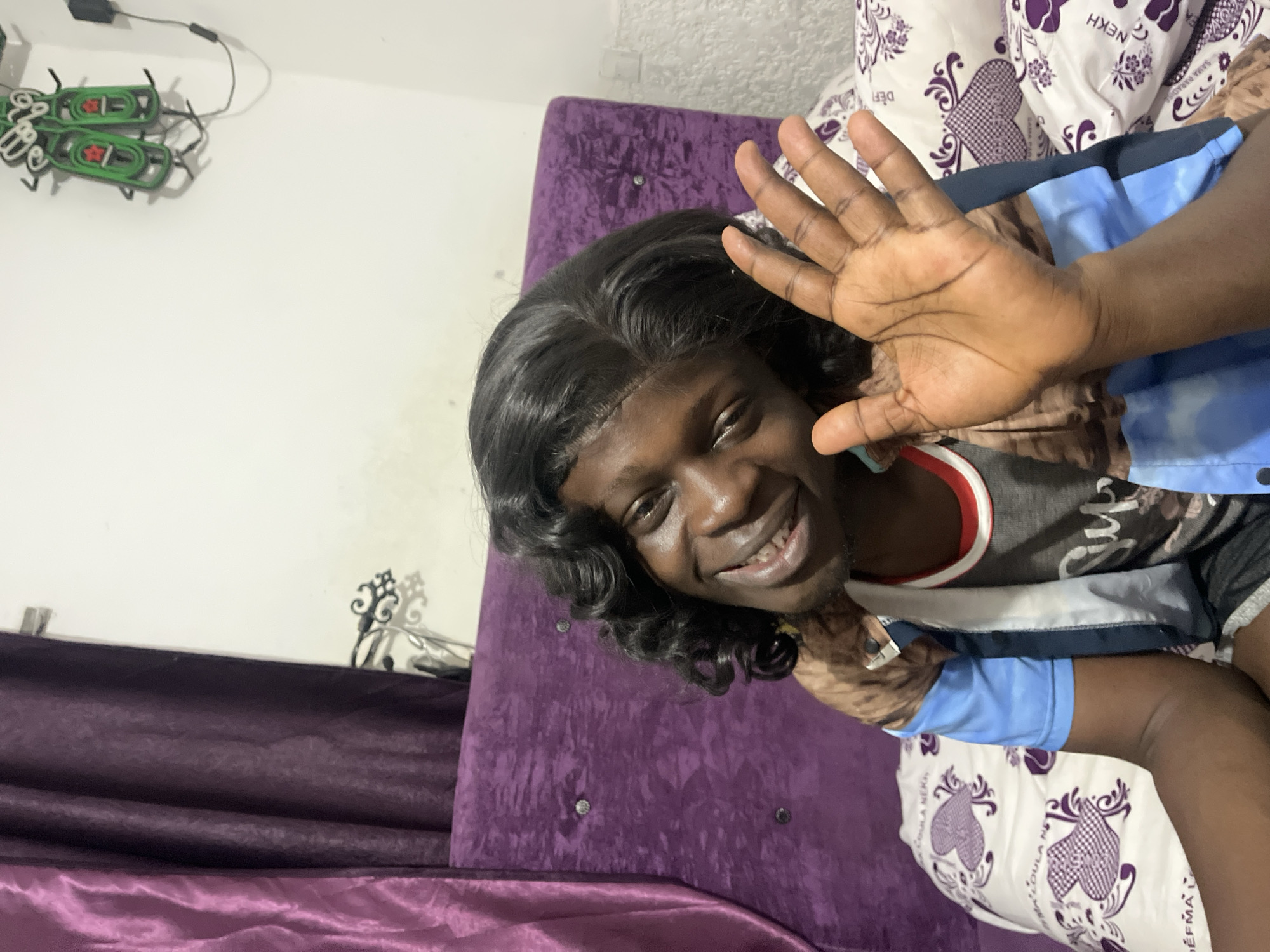 A person with short, curly black hair is shown from the chest up. The person is smiling and waving their hand in front of their face. The person is wearing a brown and blue jacket over a gray shirt with a red trim.
The person is lying on a pillow with a repeating heart pattern and the words "DEFMÃ OULA NEKH". Behind the pillow is a purple headboard. The wall behind the headboard is white. A decorative metal piece is visible on the wall next to the headboard. A green neon sign with the word "HEINEKEN" is visible on the wall near the ceiling.
Based on the information, the image cannot be conclusively identified as being in Abuja, Nigeria.