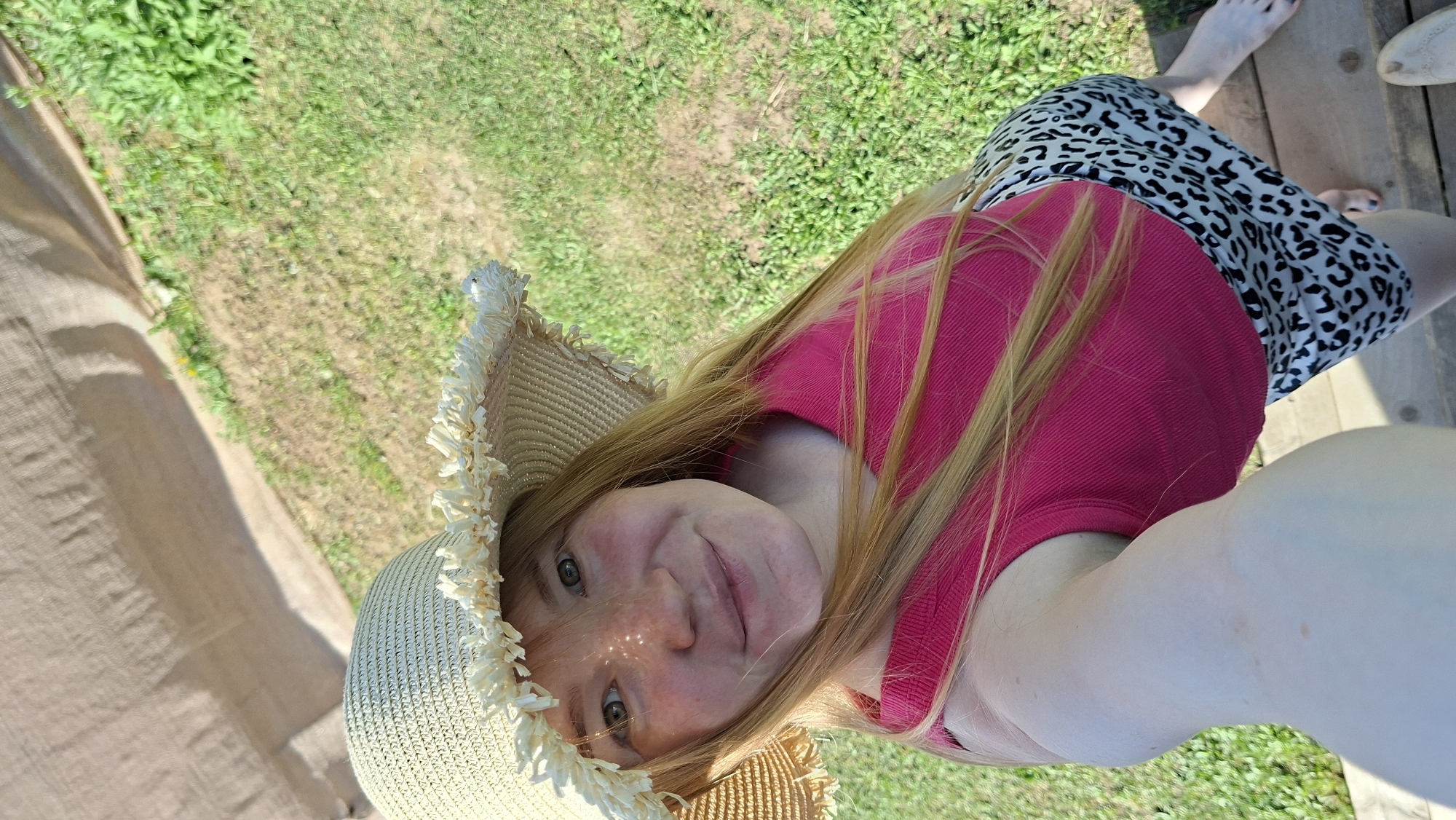 A light-skinned woman with red hair and a straw hat poses for a selfie. She is wearing a pink top and a white and black leopard-print skirt. She is standing on a wooden deck near grass, a tan canvas, and a partial view of her feet. The location is outdoors in daylight. There is no evidence to determine if the location is in Большой Букор, Russia.