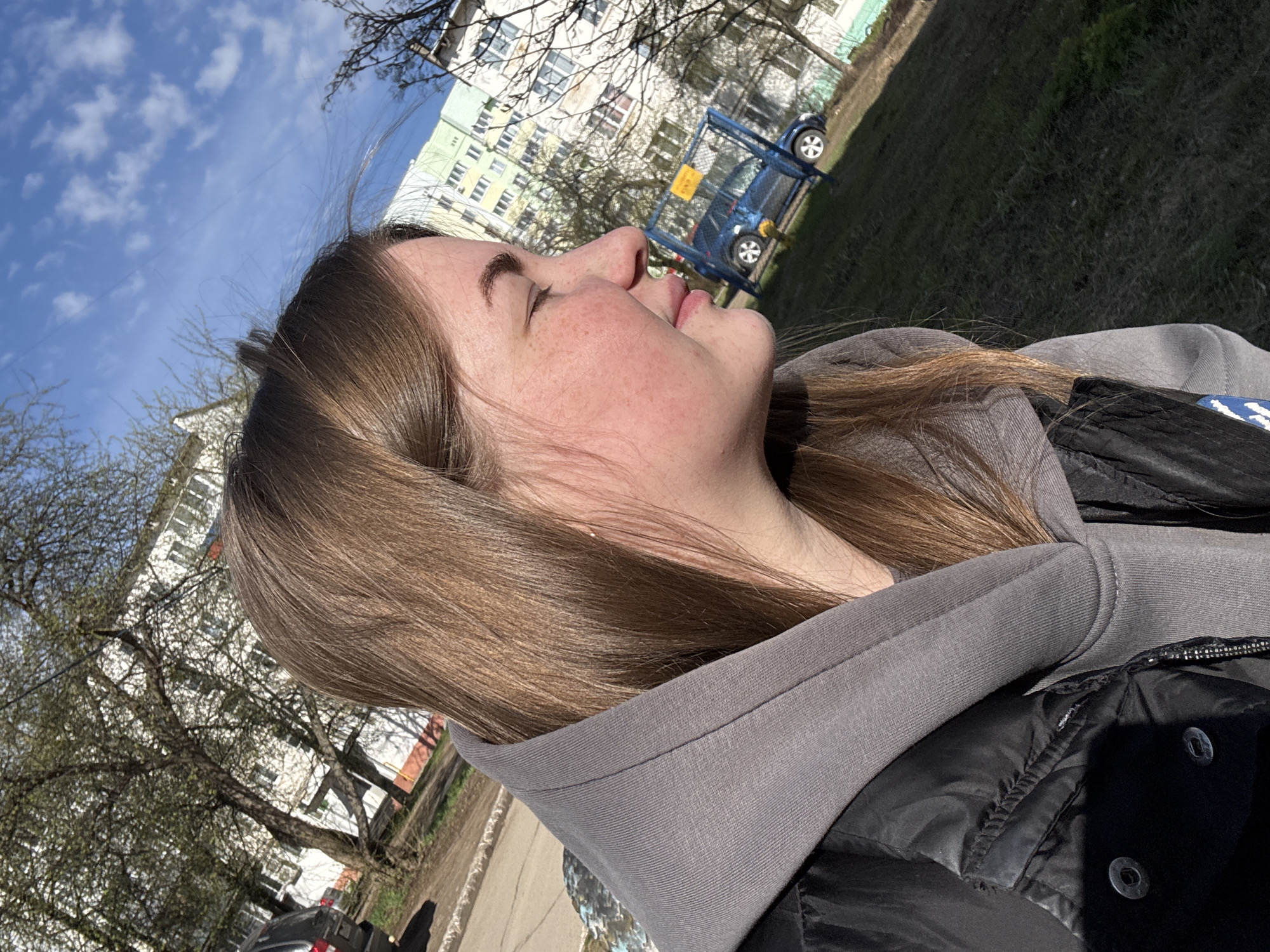 The image shows a young woman in profile, looking upwards towards a bright, partly cloudy blue sky. Her eyes are closed, and she has a calm expression, suggesting she is enjoying the sun or a moment of peace. She has light brown hair and visible freckles on her nose and cheeks.
She is wearing a grey hooded sweatshirt (hoodie) underneath a black puffer jacket.
The background depicts an urban or suburban residential area. Multi-story apartment buildings with numerous windows and light-colored facades (one light green, another white) are visible in the distance and mid-ground. Several deciduous trees, some still bare or with early spring foliage, are scattered around the buildings. To the right, there's a grassy area with a distinct blue metal structure, possibly a playground element or a small fence, and a partially visible blue car behind it. A paved surface, likely a sidewalk or road, is seen in the foreground and extending into the mid-ground.
The time of day appears to be sunny midday or early afternoon, indicated by the bright light and clear shadows. The weather is bright and clear, with scattered white clouds in the blue sky. The scene strongly suggests a typical residential courtyard or park in an Eastern European urban environment, consistent with Chaikovsky, Russia. No clearly legible text is visible within the image.