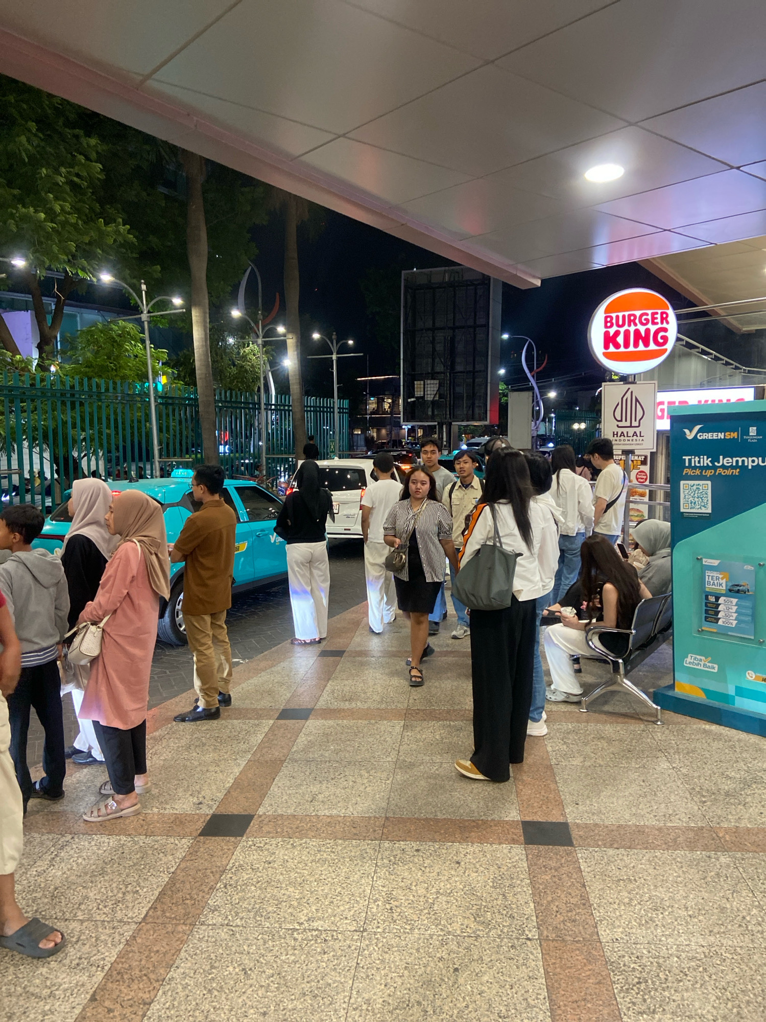 The image shows a nighttime scene outside a Burger King restaurant in Surabaya, Indonesia. A group of people are standing in a queue, some waiting to board a turquoise taxi. The Burger King sign is illuminated above the entrance. There are streetlights and trees visible in the background. The ground is paved with tiles.